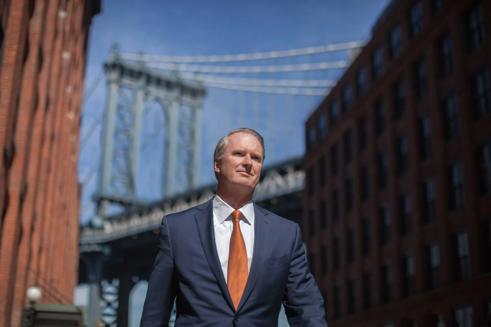 A man in a suit with an orange tie standing outdoors in front of a cityscape that includes the Brooklyn Bridge and brick buildings.