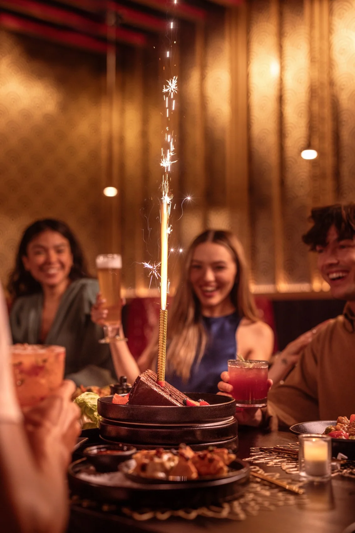 People celebrating at a dinner party with a birthday cake topped with a sparking birthday candle, smiling and holding drinks in a warmly lit room.