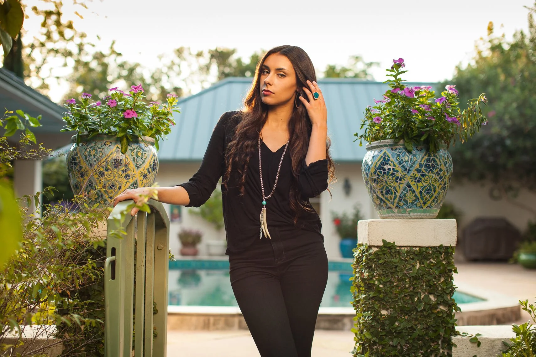A woman dressed in black standing outdoors near a pool with large decorative flower pots on either side, during golden hour with trees in the background.
