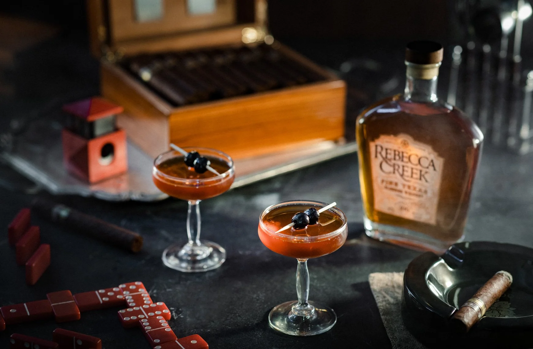 Two Tom Collins cocktails garnished with blueberries and a cherry on a black surface with a bottle of Rebecca Creek whiskey, a cigar, dominoes, a cigar holder, a cigar cutter, and a game box in the background.
