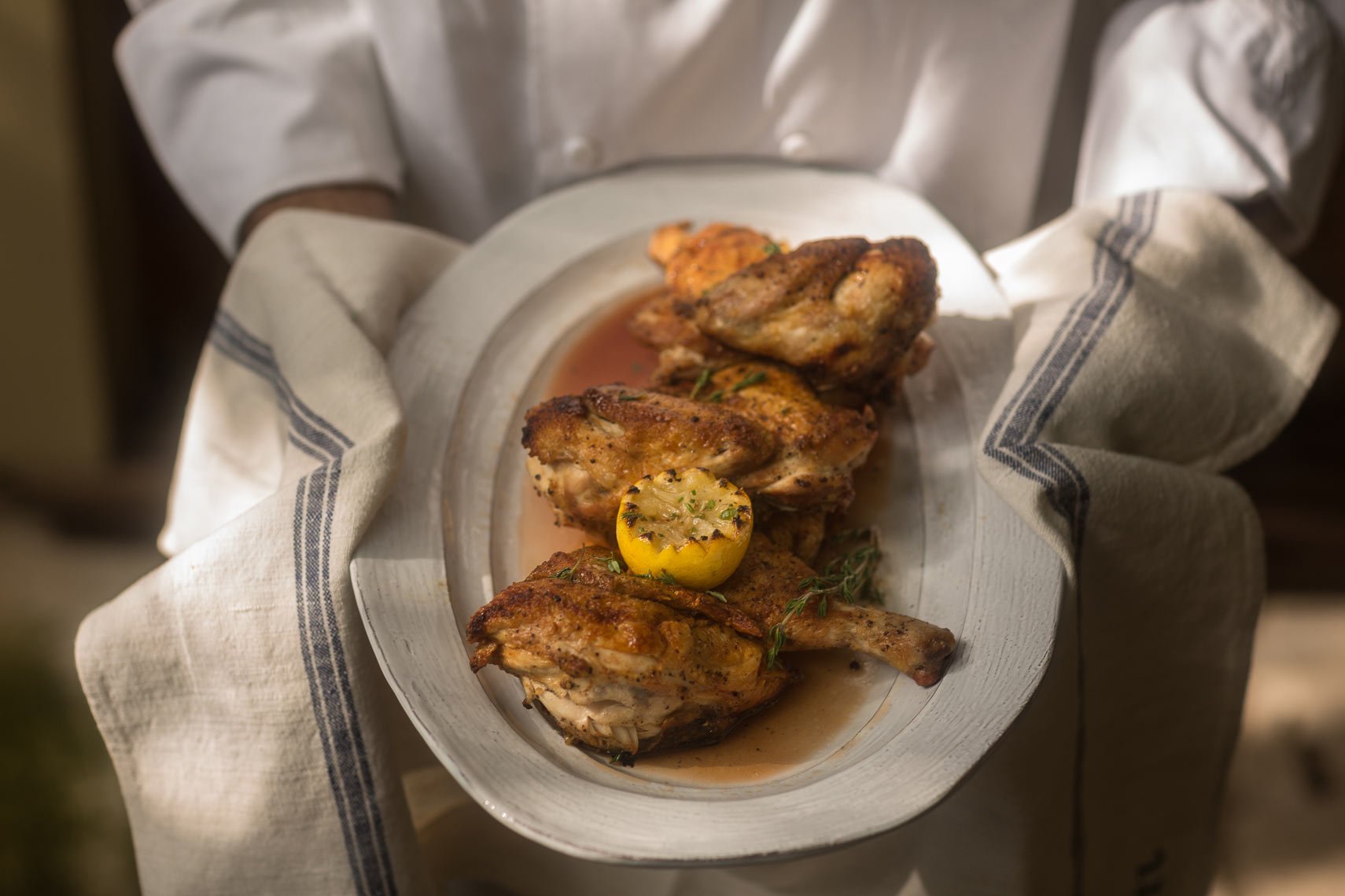 Person holding a white platter with grilled chicken quarters garnished with herbs and sliced lemon.