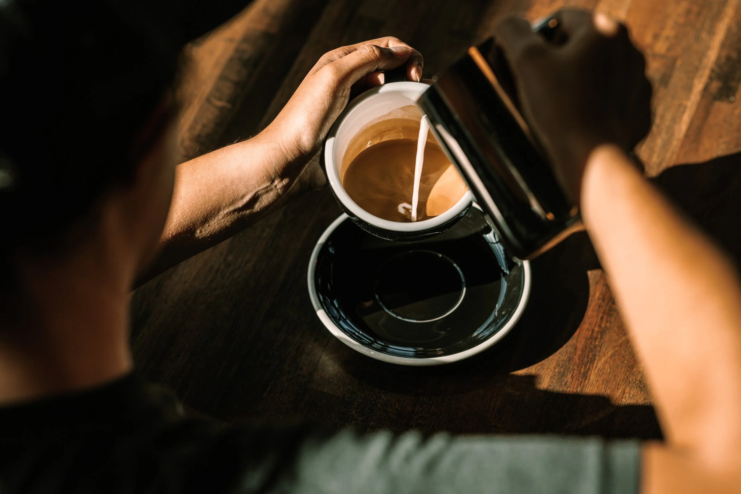 A person pouring cream into a cup of coffee on a wooden table.