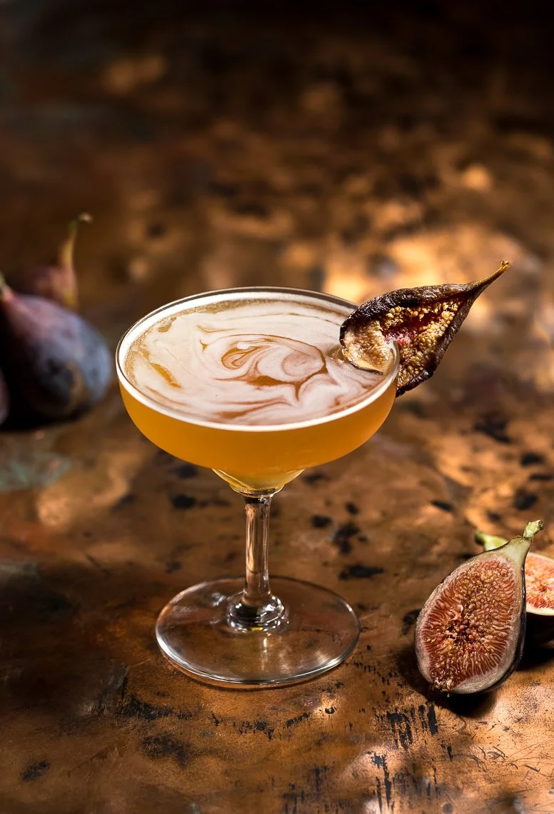 A cocktail in a glass garnished with a dried fig, placed on a rustic wooden surface with fresh figs nearby.