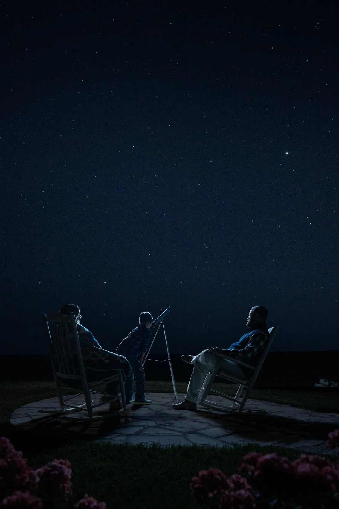 A family of three sitting outside at night, looking at the stars. The sky is clear and filled with stars, and one child is pointing with a telescope toward the sky.