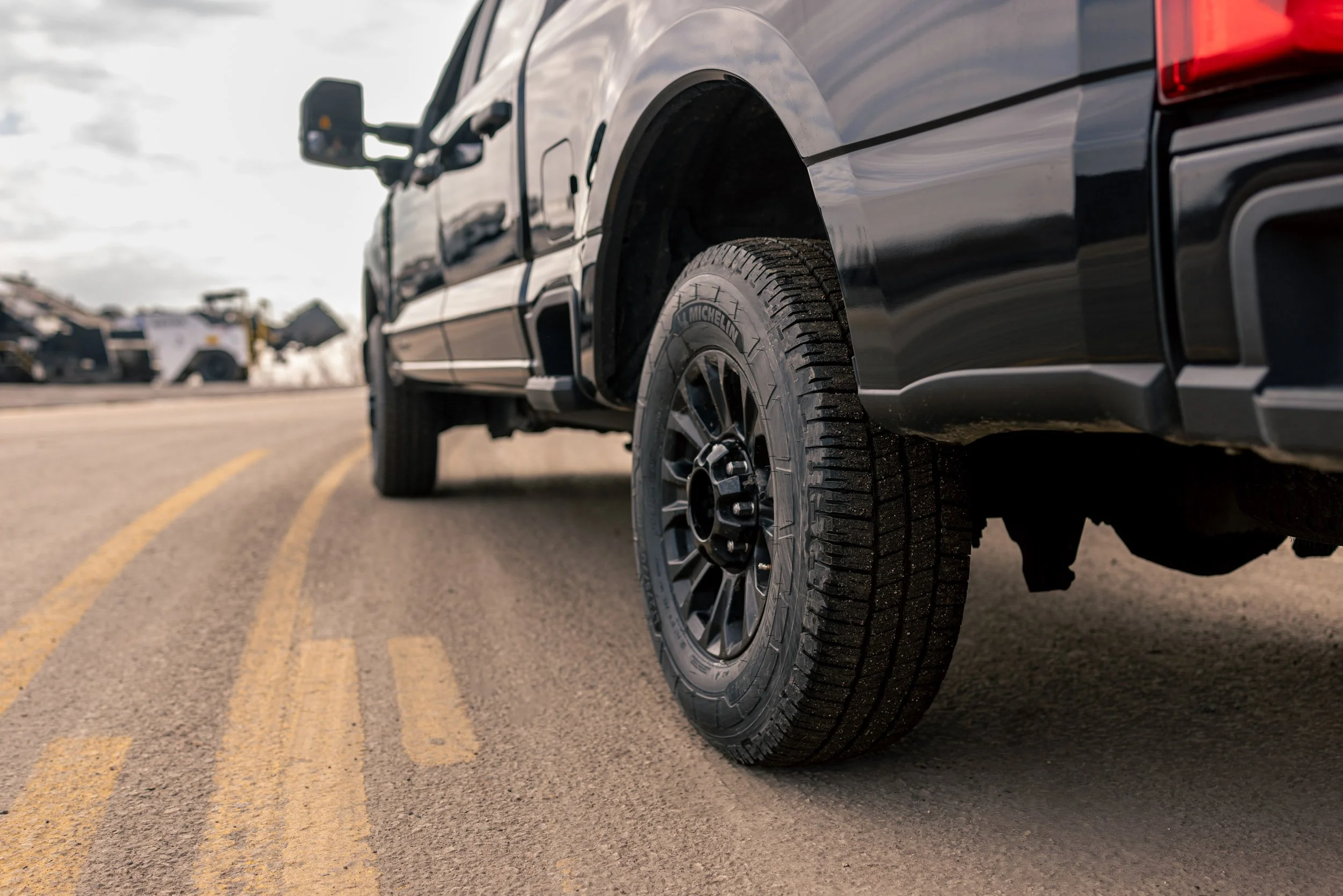 Close-up of a black pickup truck's tire and wheel on a paved road with yellow lines, with a blurred background of trucks and cloudy sky.