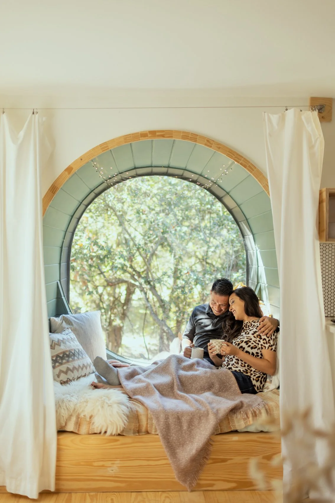 A couple sits on a bed inside a cozy room with a large circular window, enjoying each other's company and looking at a phone together.