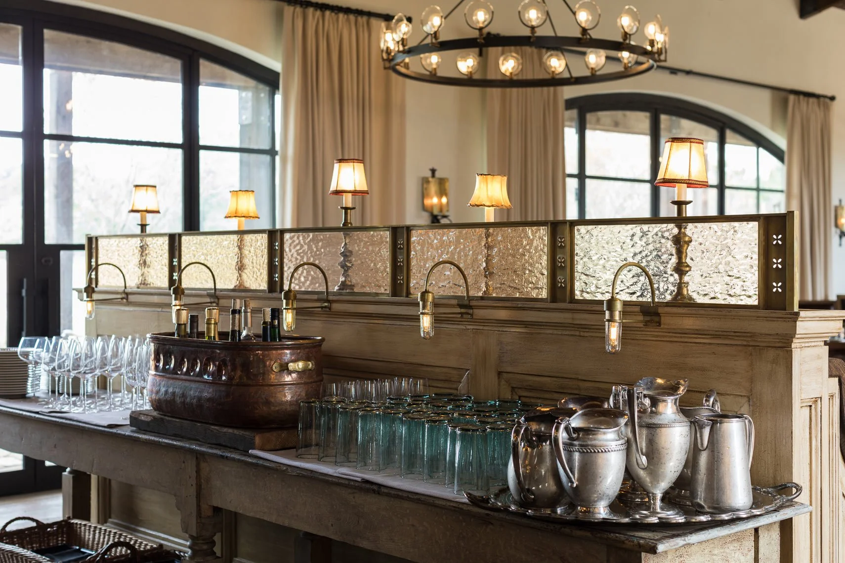 A bar setup with a wooden counter, glassware, and silver pitchers. Behind the counter is a divider with textured glass panels and mounted vintage-style lamps. The background features large windows with curtains and a large circular chandelier with ex