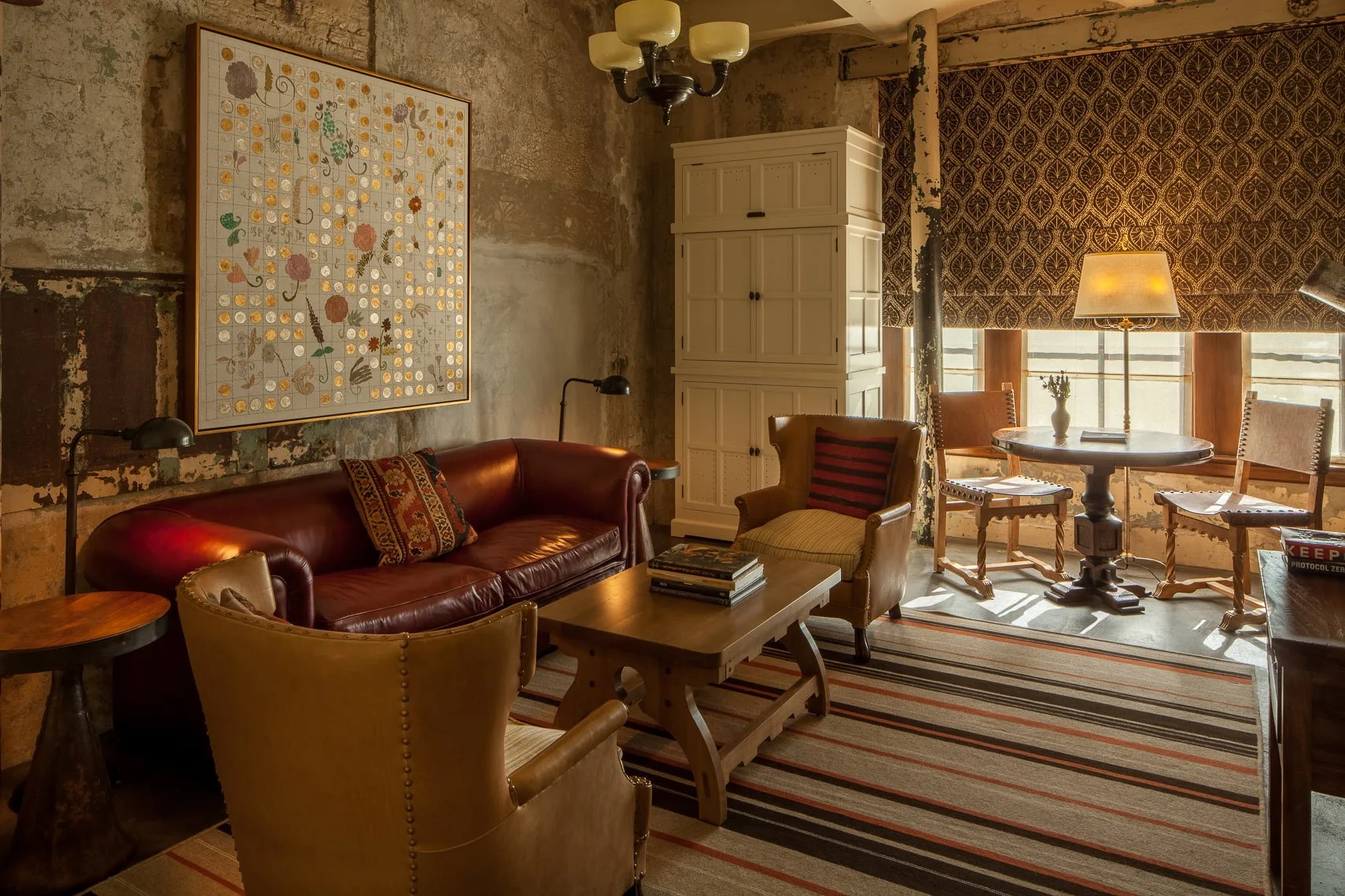 Cozy living room with a red leather sofa, wooden coffee table, vintage armchair, side table, striped area rug, large window with patterned curtains, and a mix of rustic and vintage decor, including a wall painting and a white cabinet.