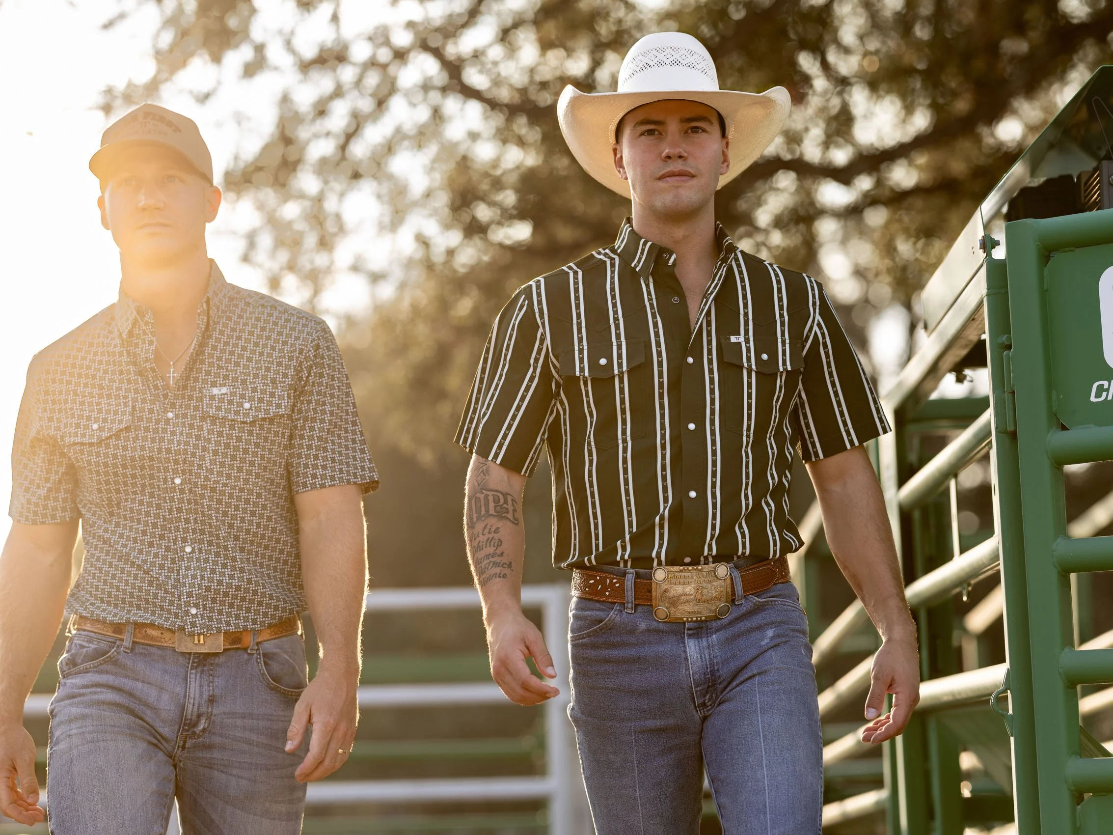 Two men walking outdoors, one wearing a white cowboy hat and a striped black and white shirt, the other in a patterned shirt and a baseball cap, with a green metal railing on the right and blurred tree leaves in the background, during sunset.