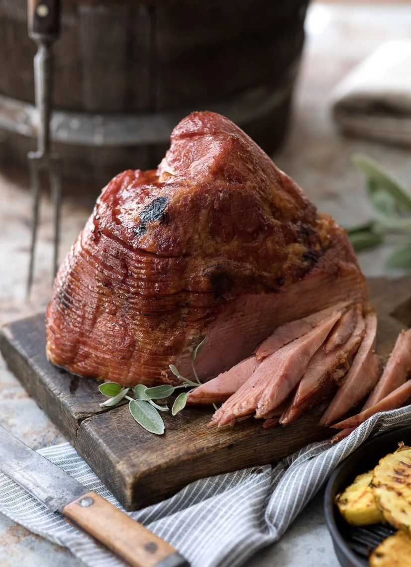 A large, cooked ham with a portion sliced, placed on a wooden cutting board with some sprigs of fresh herbs, set on a striped cloth on a rustic table.