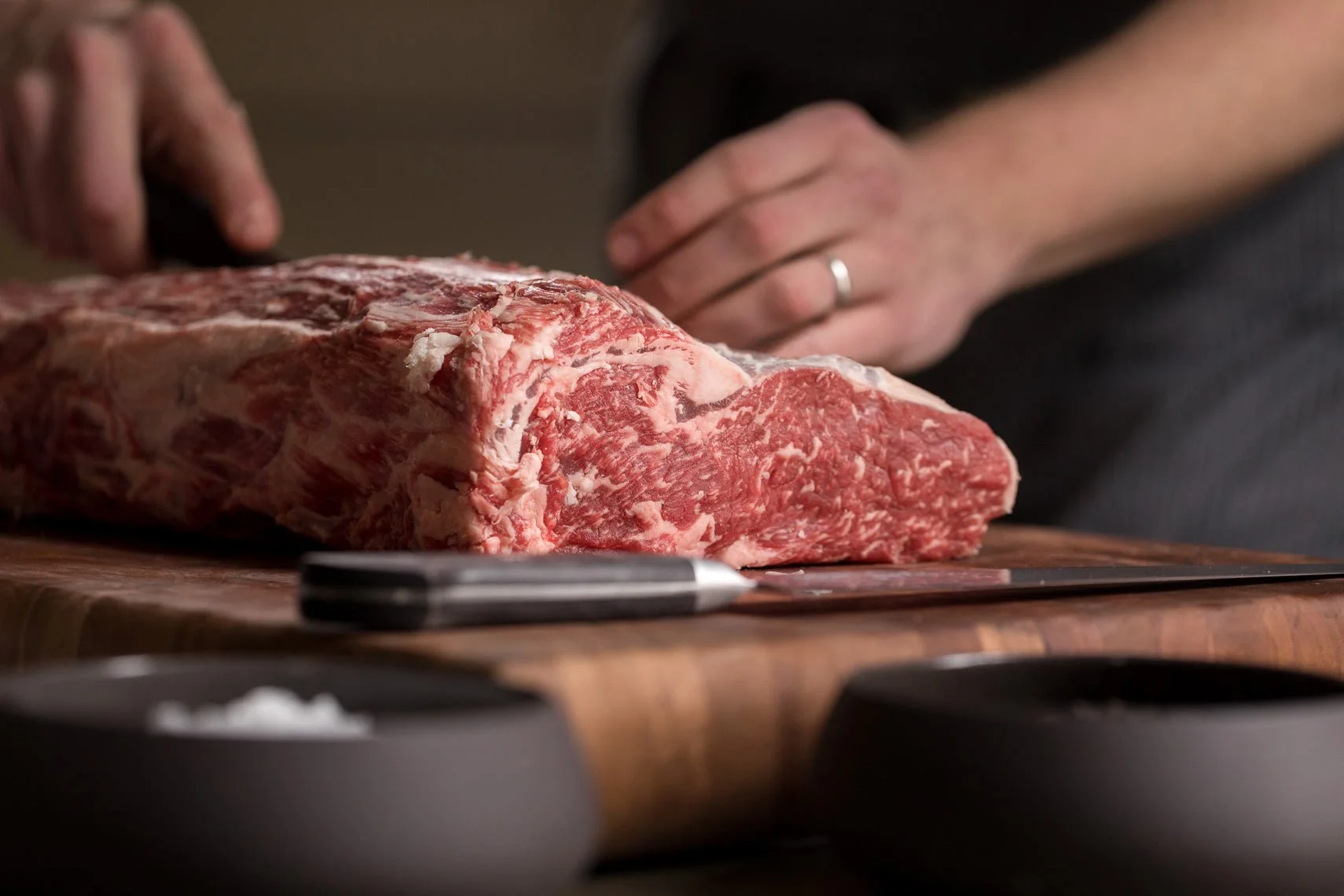 A person is preparing a large cut of marbled raw beef steak on a wooden cutting board with a knife nearby.