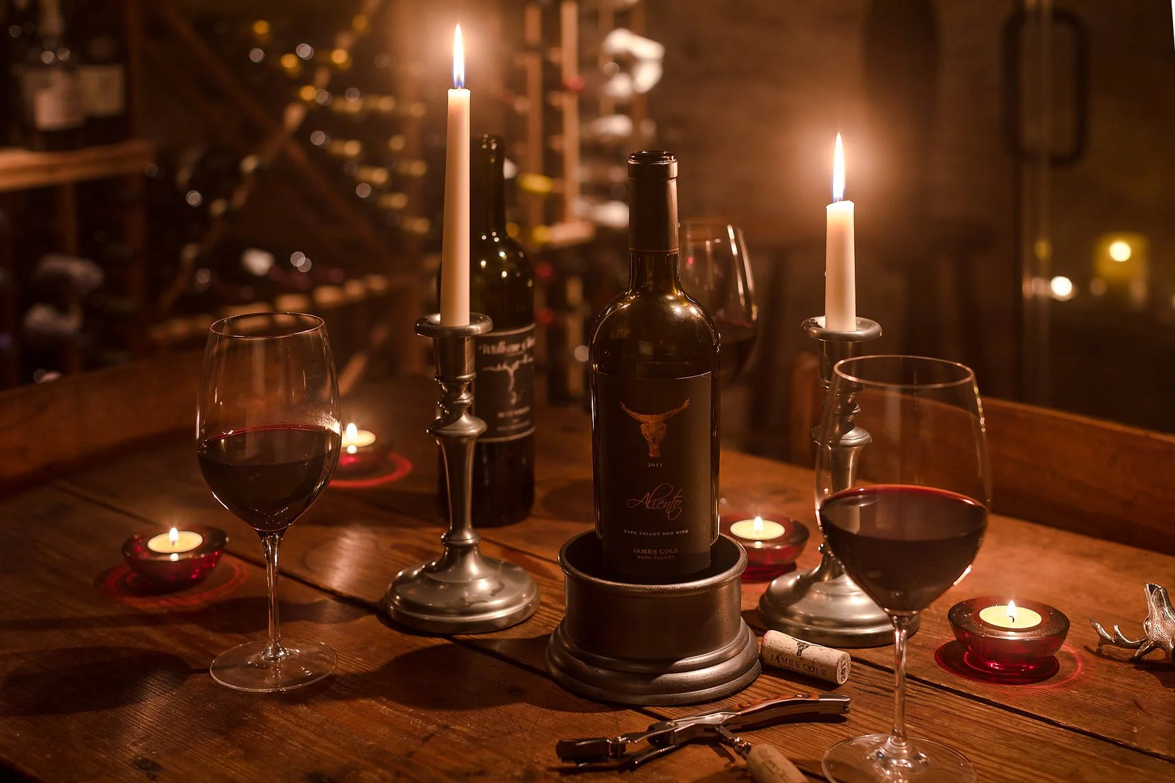 A romantic dinner setup with two glasses of red wine, lit candles, wine bottles, and a cork on a wooden table in a warm, cozy setting.