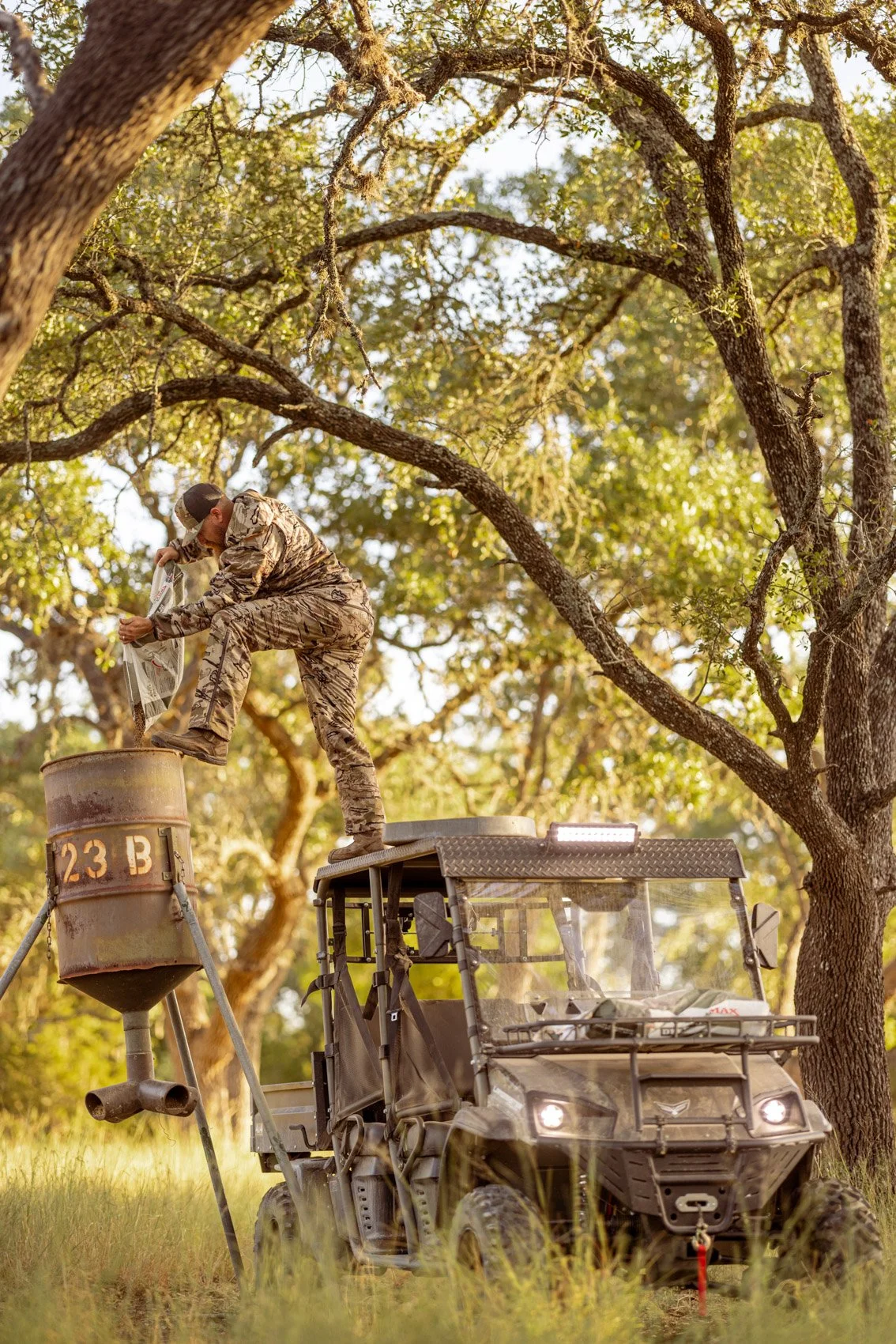A person in camouflage clothing gathers or pours seeds or grains from a large metal container onto a cylindrical feeder, which is mounted on a tripod. The person is standing on a vehicle platform next to the feeder. The scene takes place outdoors in 