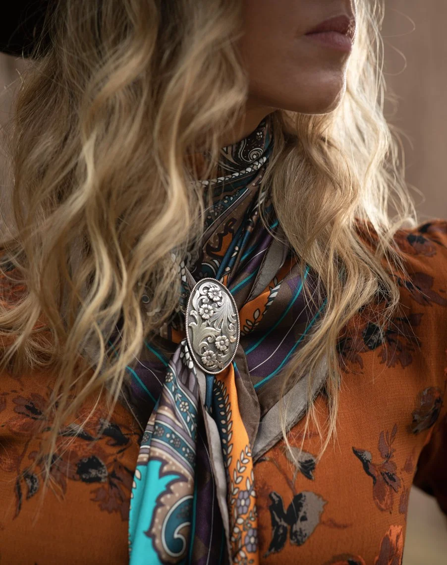 Close-up of a woman with wavy blonde hair wearing a patterned scarf with floral design and a large silver brooch, dressed in an orange floral blouse.