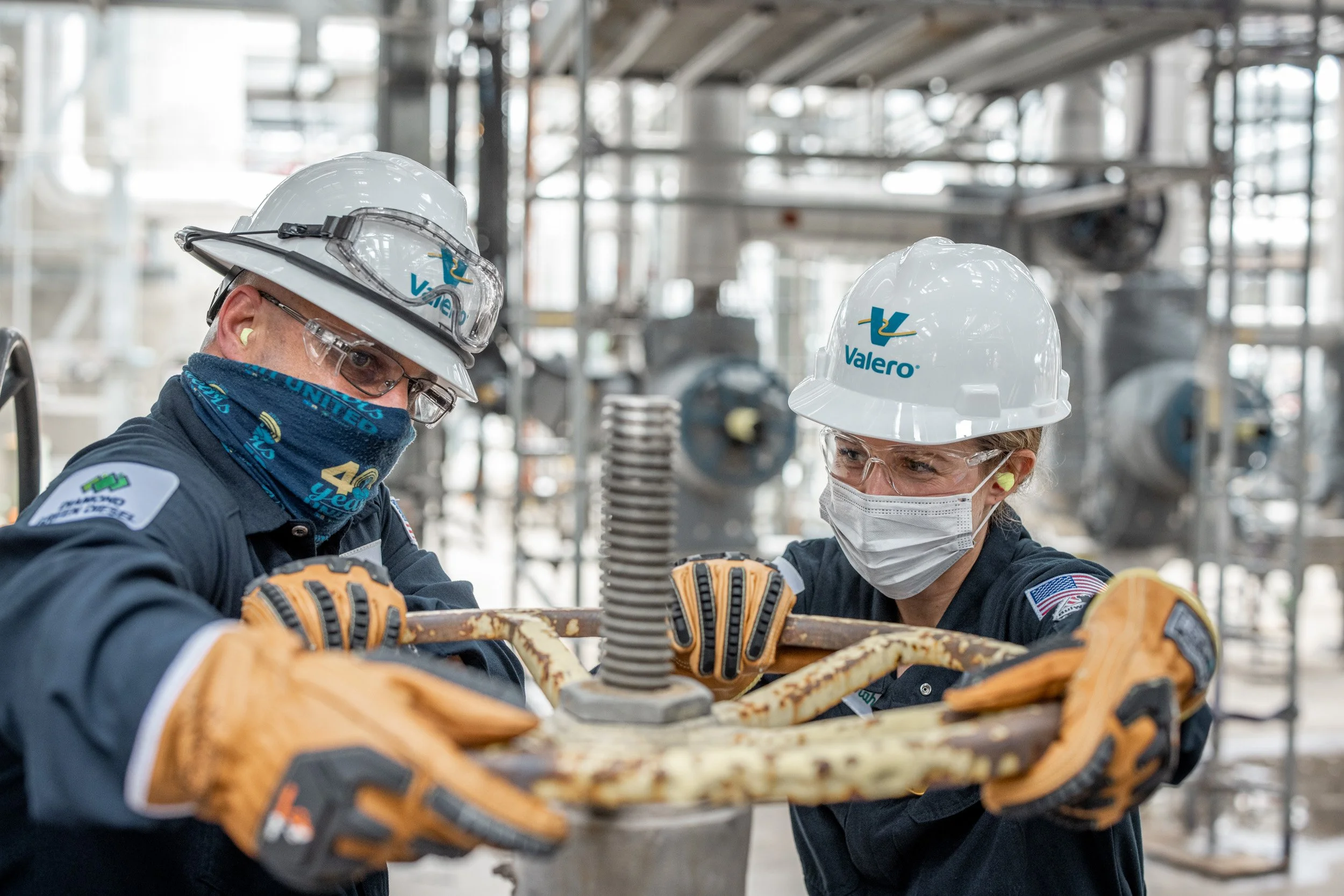 Two workers wearing hard hats, safety glasses, masks, and gloves working together on a mechanical task in an industrial setting, with machinery and equipment in the background.