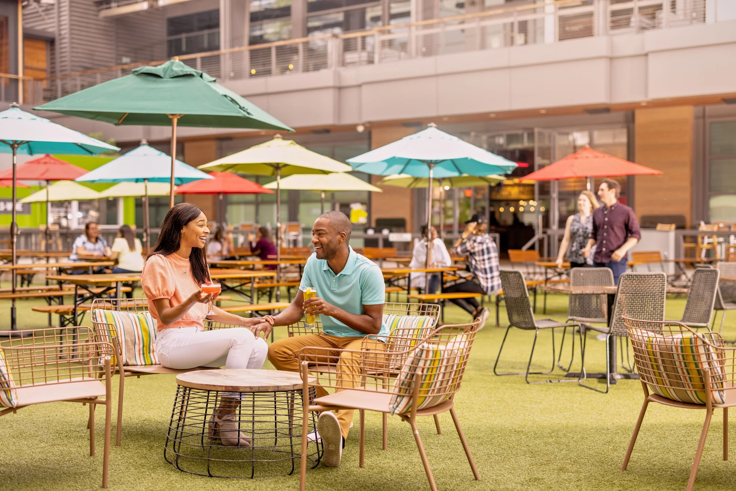 A man and woman sitting at an outdoor café table, holding hands and smiling at each other, with colorful umbrellas in the background.