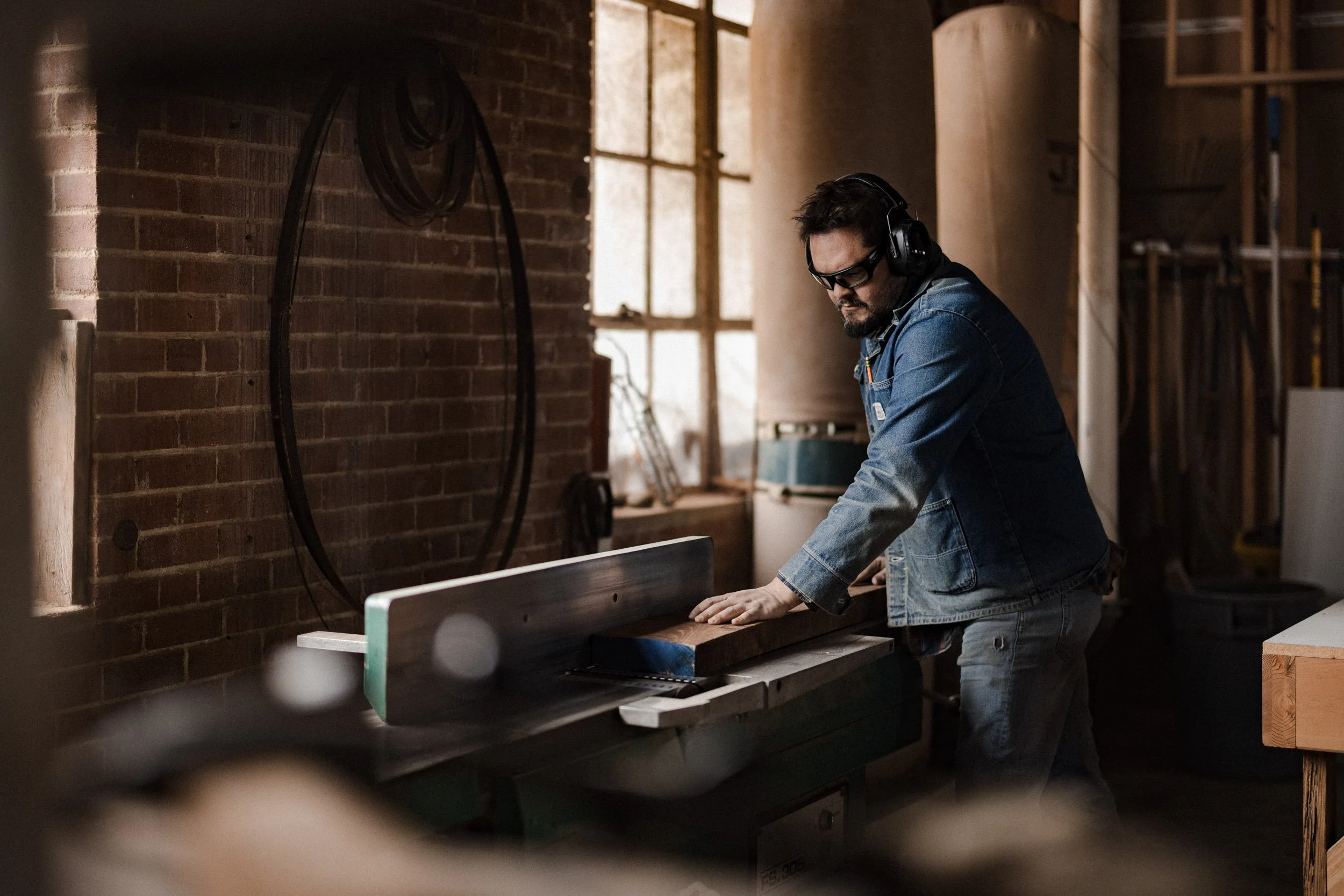 A man wearing safety goggles, headphones, and a denim jacket is working in a woodworking shop, leaning over a workbench.