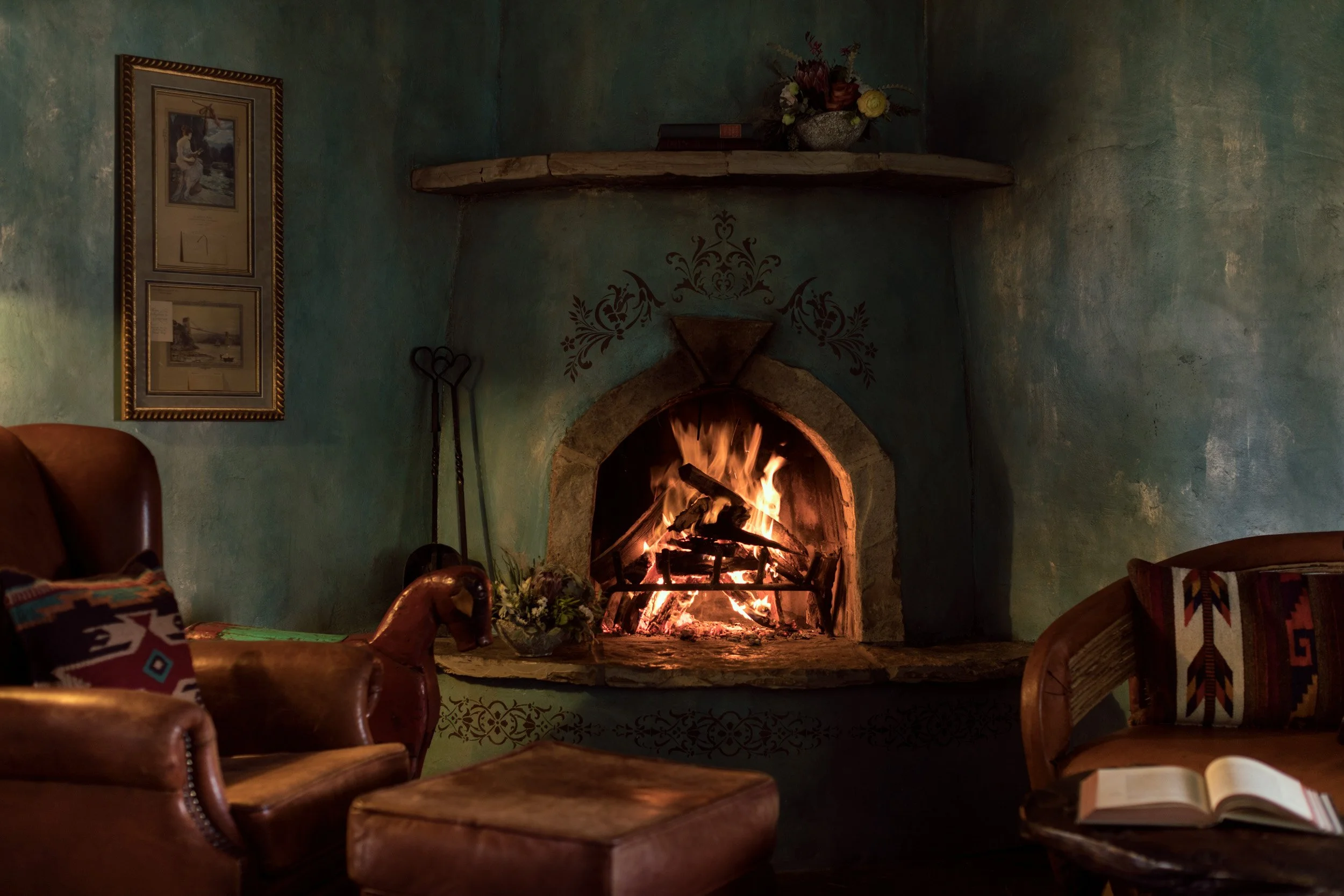 A cozy living room with a lit fireplace, surrounded by wooden chairs, a framed picture on the wall, and decorative items including a bouquet of flowers on the mantle.
