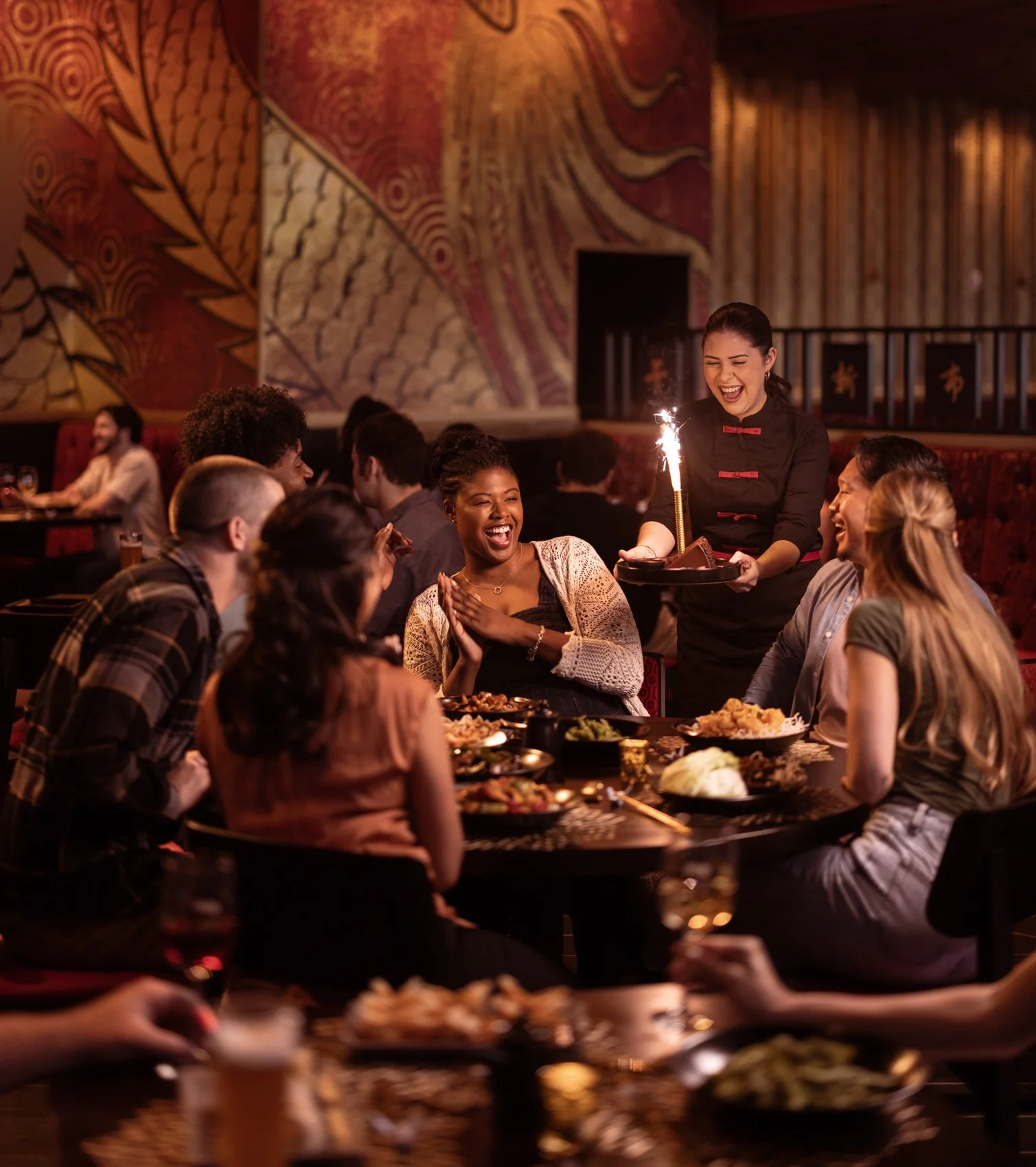Group of diverse friends celebrating with a meal at a restaurant, with a woman holding a lit sparkler and serving food, others smiling and enjoying their time.