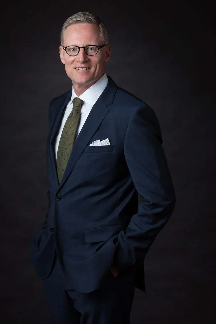 A man in a dark blue suit, white shirt, and green tie poses against a black background.