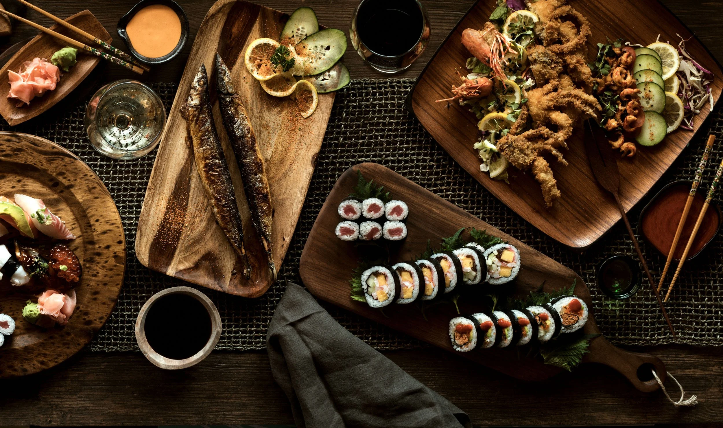 Assorted Japanese dishes on wooden trays, including sushi rolls, grilled fish, sashimi, salad, and dipping sauces, arranged on a woven tablecloth.