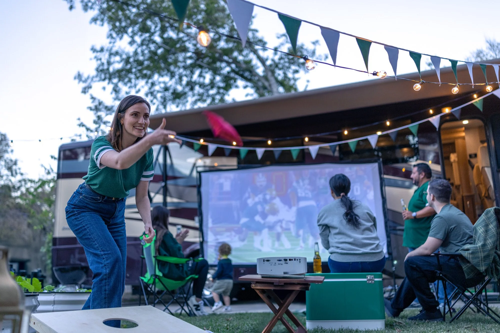 People gathered outdoors for a movie night, with a large screen showing a film, string lights and banners decorating the area, and a woman in a green shirt playing a game or rotating an object in the foreground.