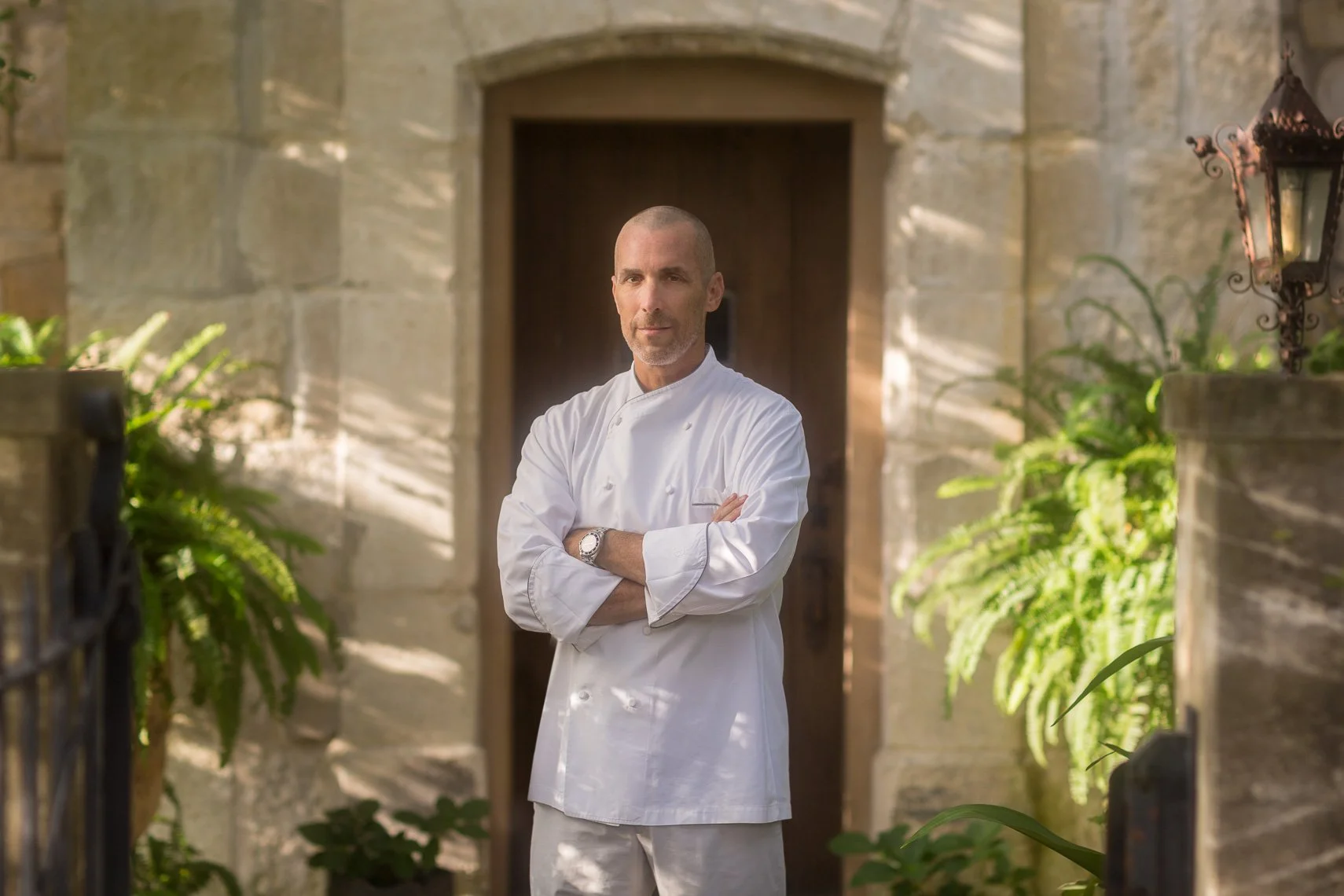 A male chef in a white uniform standing outdoors with arms crossed, in front of a stone building with a wooden door, surrounded by greenery.