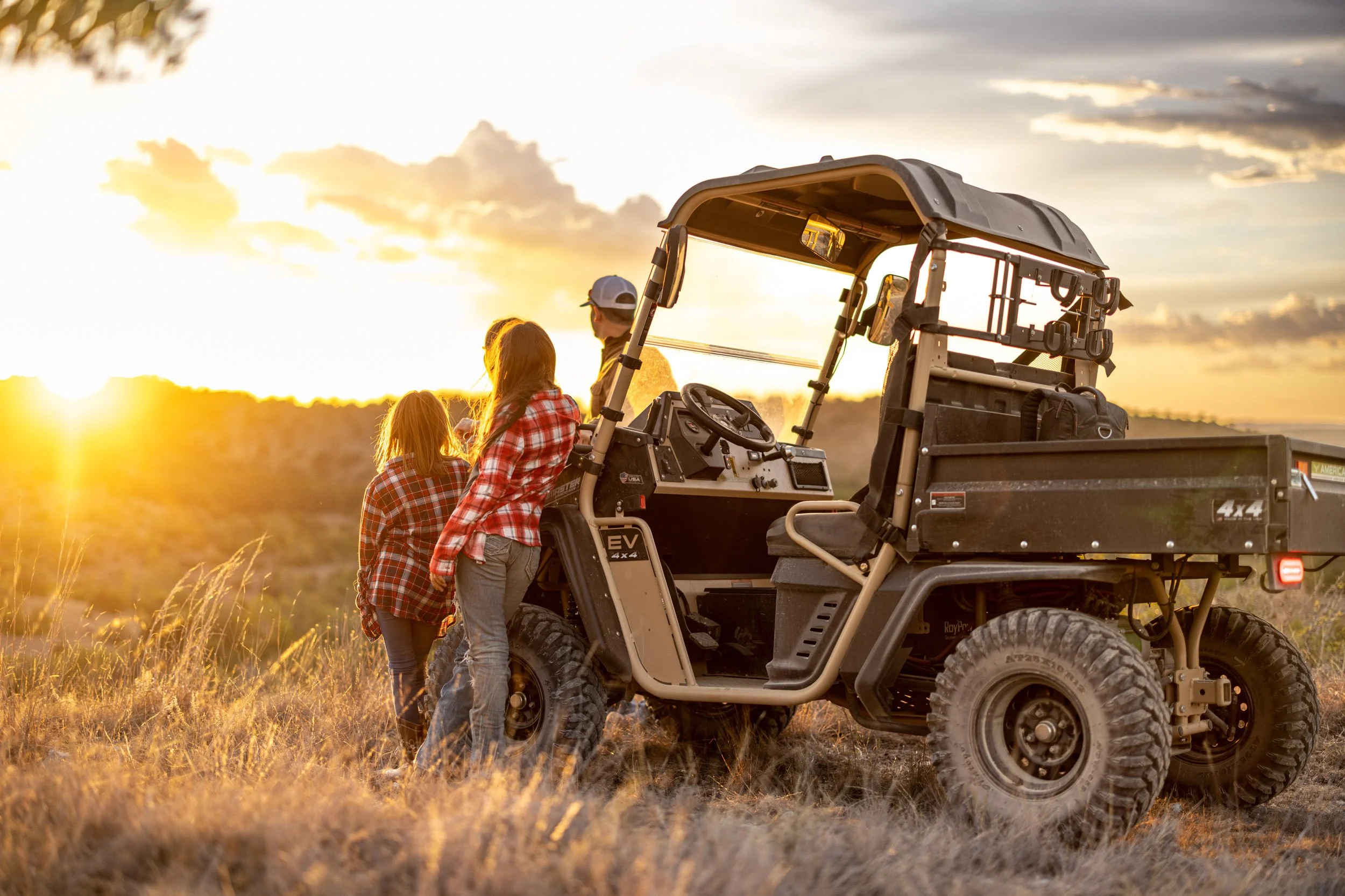 Three people, wearing plaid shirts and jeans, stand beside a utility vehicle in a field during sunset. The vehicle is off-road with a canopy, and the sky is partly cloudy with warm lighting.