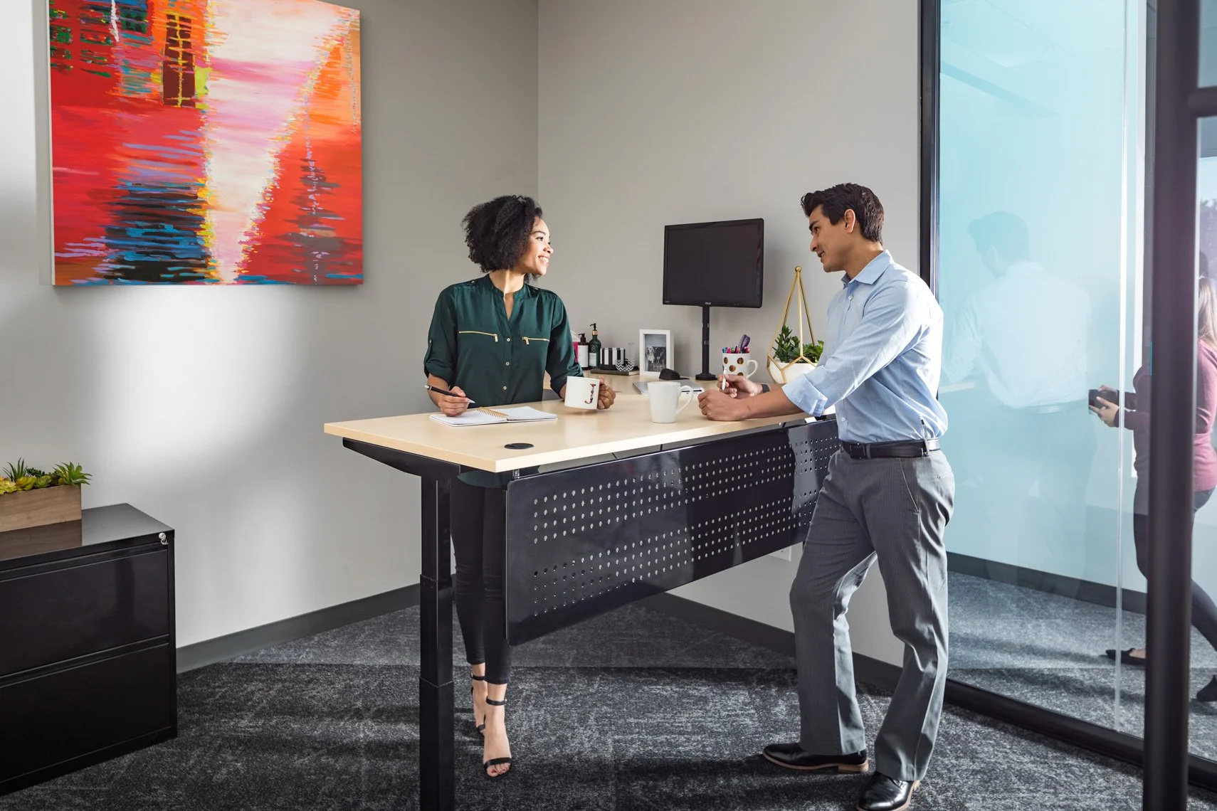 A man and woman in business attire are smiling and engaged in conversation in a modern office. The woman is standing behind a desk with a notebook and coffee mug, while the man is leaning on the desk holding a cup. There is a colorful abstract painti