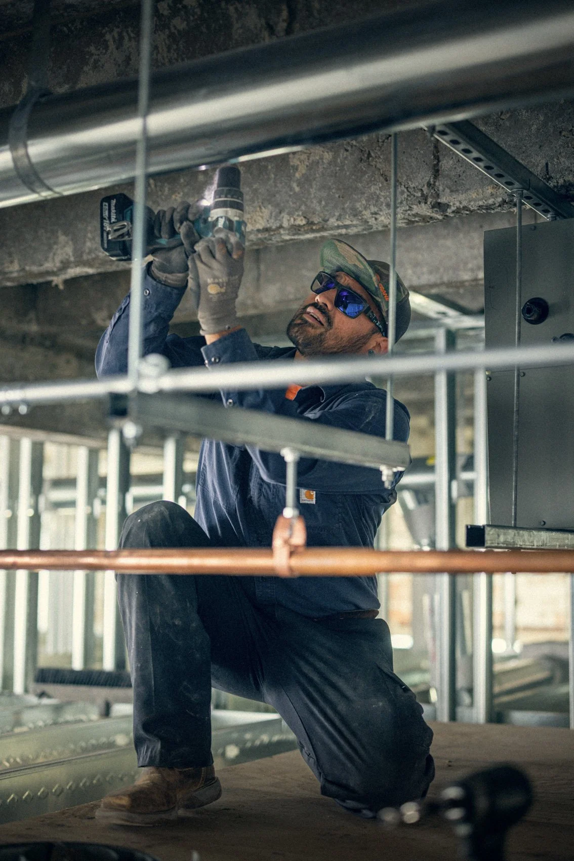 A construction worker wearing safety glasses, gloves, a hat, and work clothes is kneeling and using a power drill on a metal pipe under a concrete ceiling.