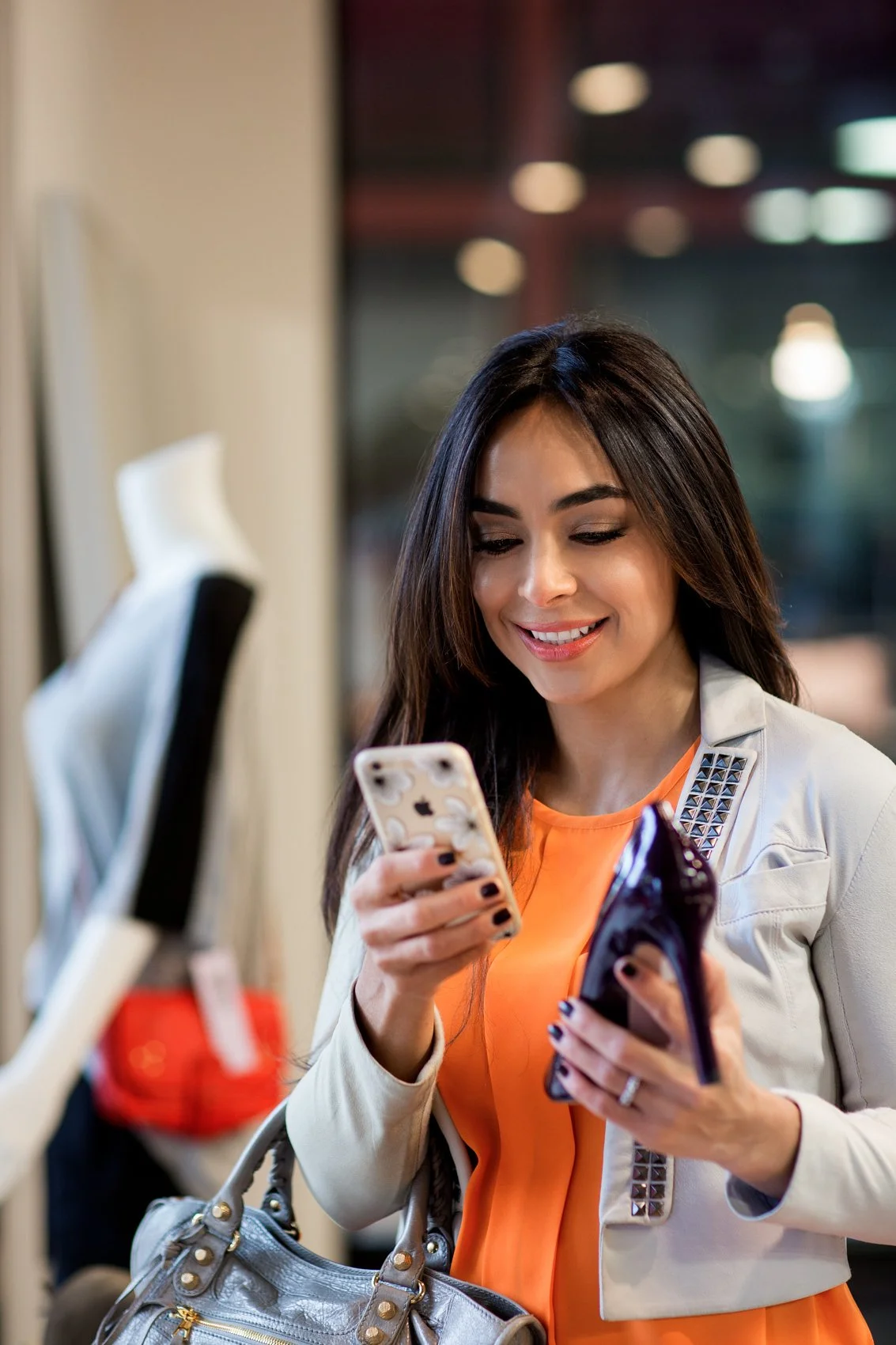 A woman with long dark hair, wearing an orange top and light-colored jacket with studded details, smiling while looking at her smartphone in a retail store.