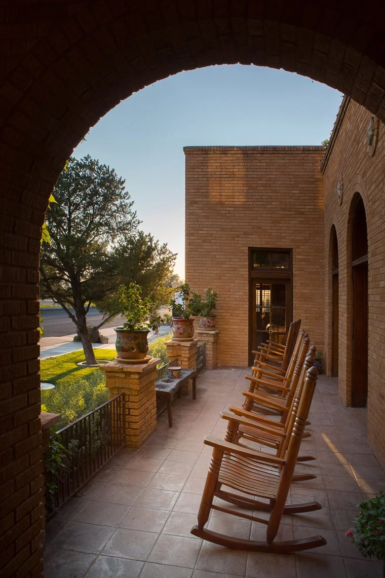 A covered porch with a row of wooden rocking chairs facing outward, brick walls, potted plants, and a tree in the background at sunset.