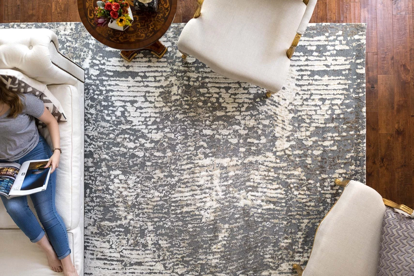 Top-down view of a living room with a patterned grey and white rug. A woman is lying on a white sofa, reading a magazine. There is a round wooden table with a floral arrangement in the center, and two upholstered chairs with gold accents.