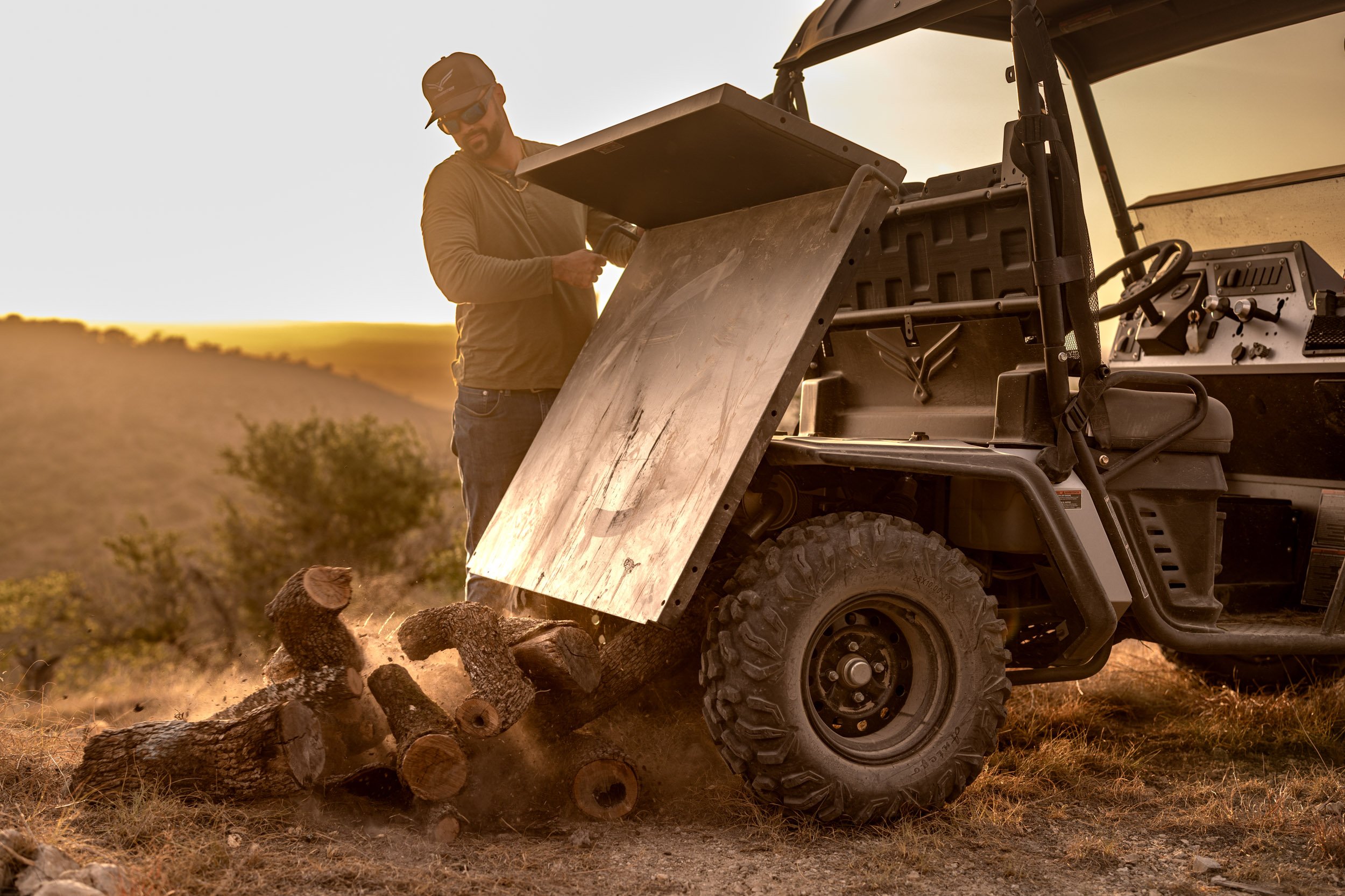 A man wearing sunglasses and a cap is operating a utility vehicle with a metal dump bed, loading logs and small wood pieces in a rural outdoor setting during sunset.