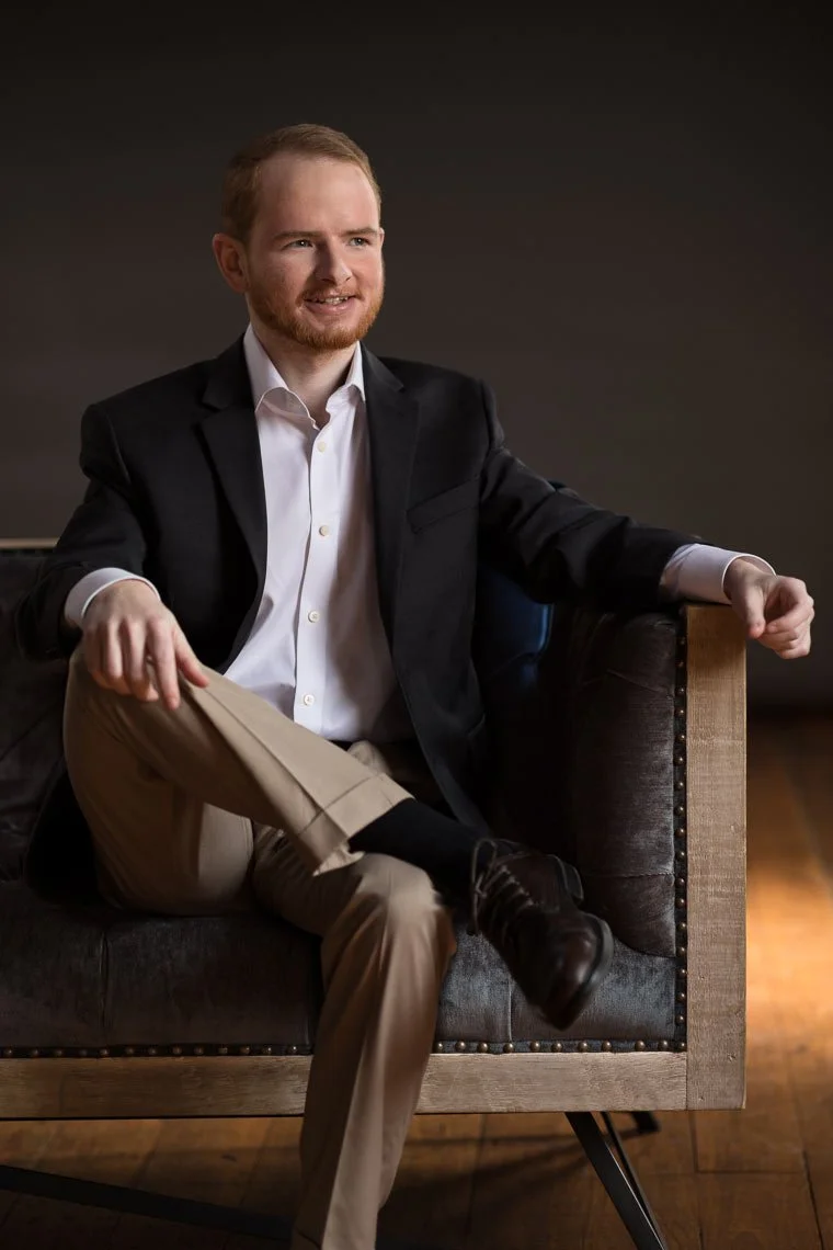 A young man with red hair and a beard sitting on a dark sofa, wearing a black blazer, white shirt, khaki pants, black socks, and brown shoes, posing with a confident smile in a dimly lit room.