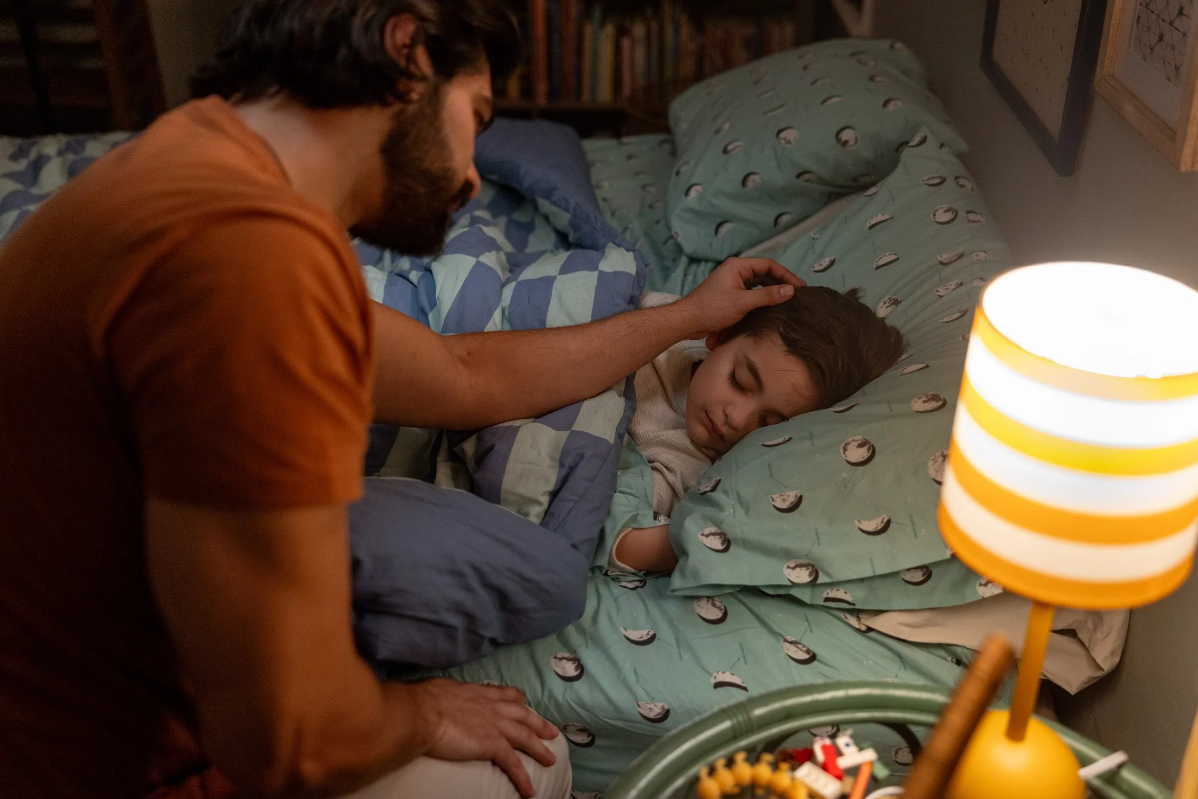 A man with dark hair and beard wearing an orange shirt is comforting a young girl with long brown hair who is sleeping peacefully on a bed with patterned blue and green bedding. The bed has pillows and a bookcase appears in the background. A small ye