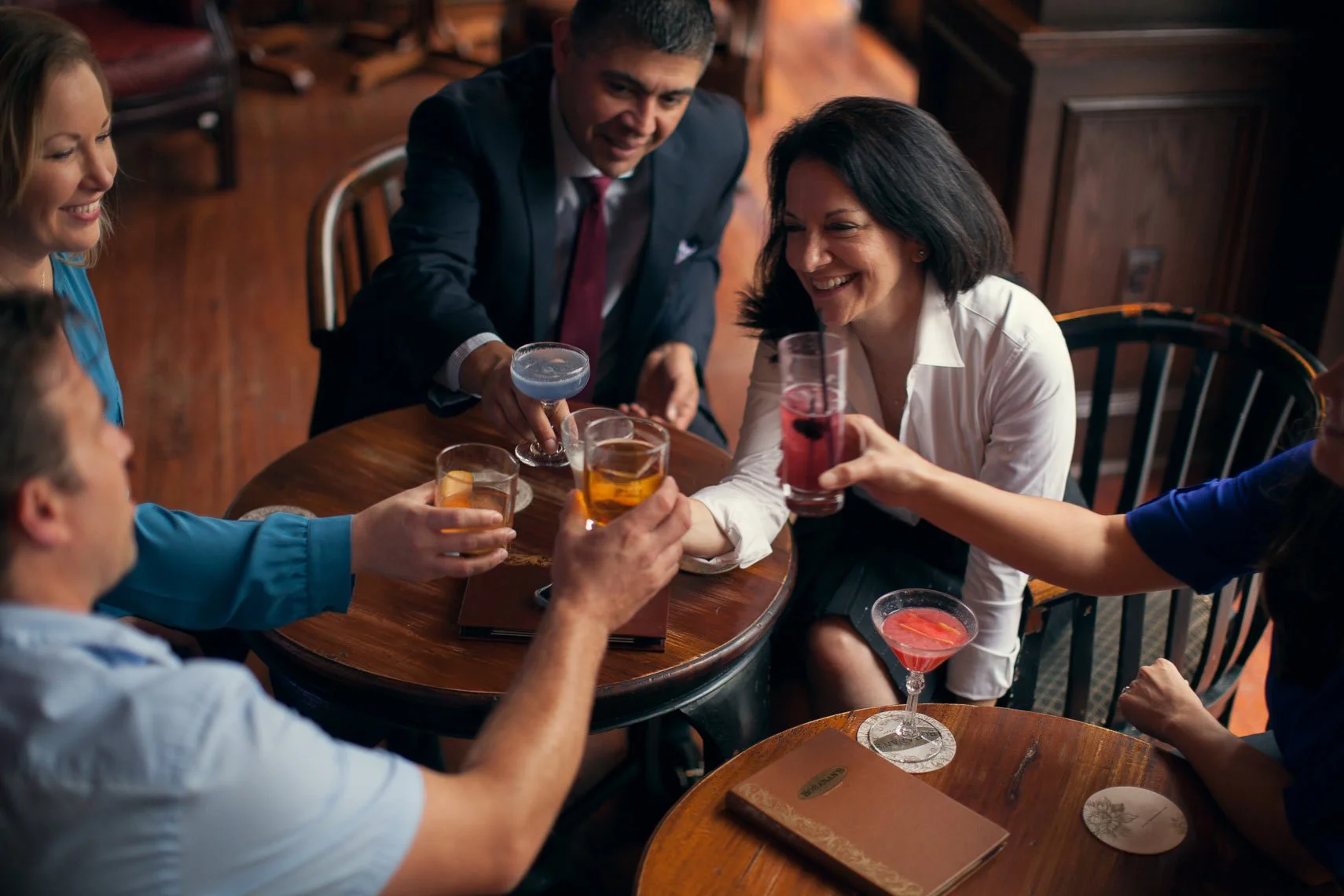 Group of friends enjoying drinks and toasting around a wooden table in a bar or restaurant.
