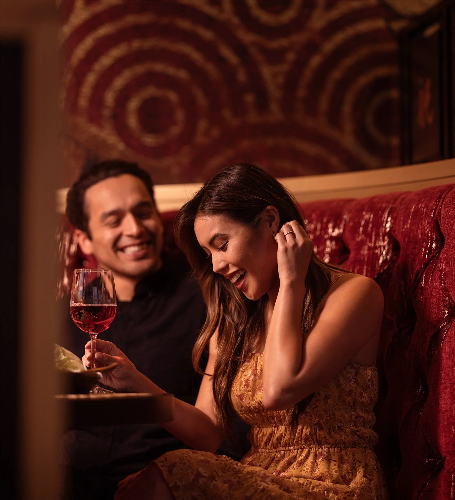 A man and a woman sitting in a dimly lit restaurant or bar, smiling and laughing. The woman is holding a glass of red wine, and the man is looking at her, appearing happy. The background has a decorative wall with circular patterns.