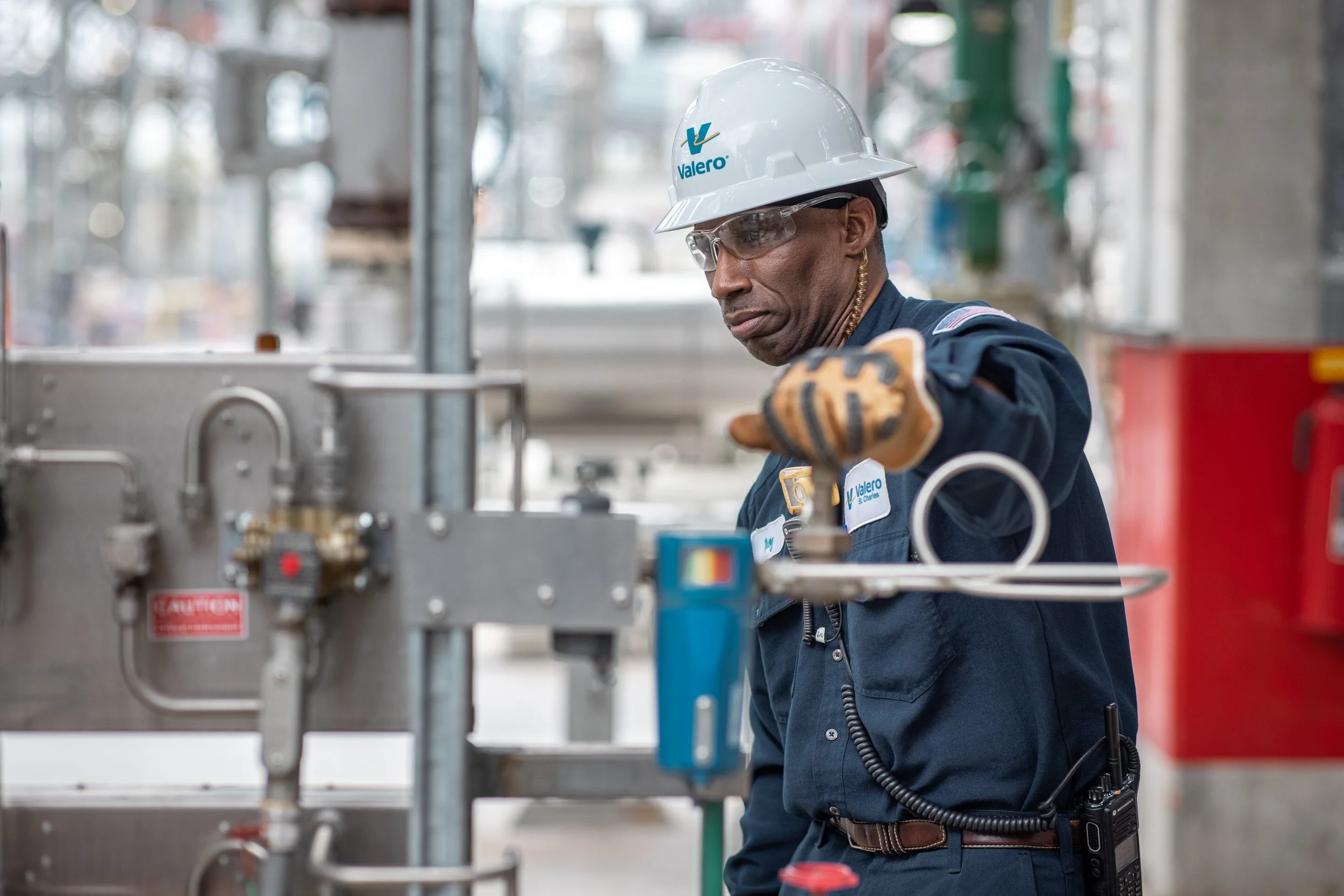 A worker in safety gear, including a Valero helmet and safety glasses, inspects industrial equipment at an industrial facility.