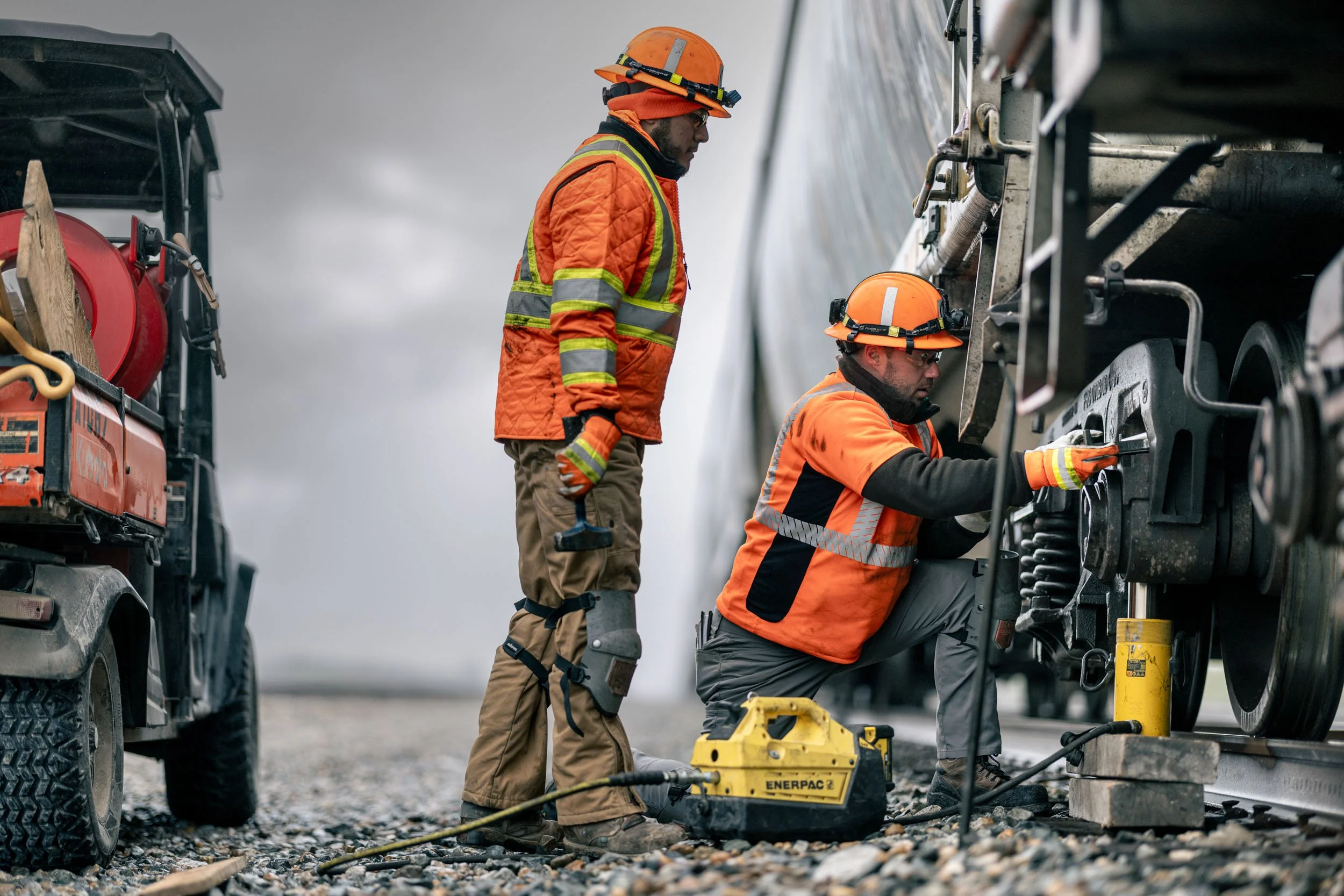 Two construction workers in orange safety jackets and hard hats repair a large piece of industrial equipment outdoors on a cloudy day.