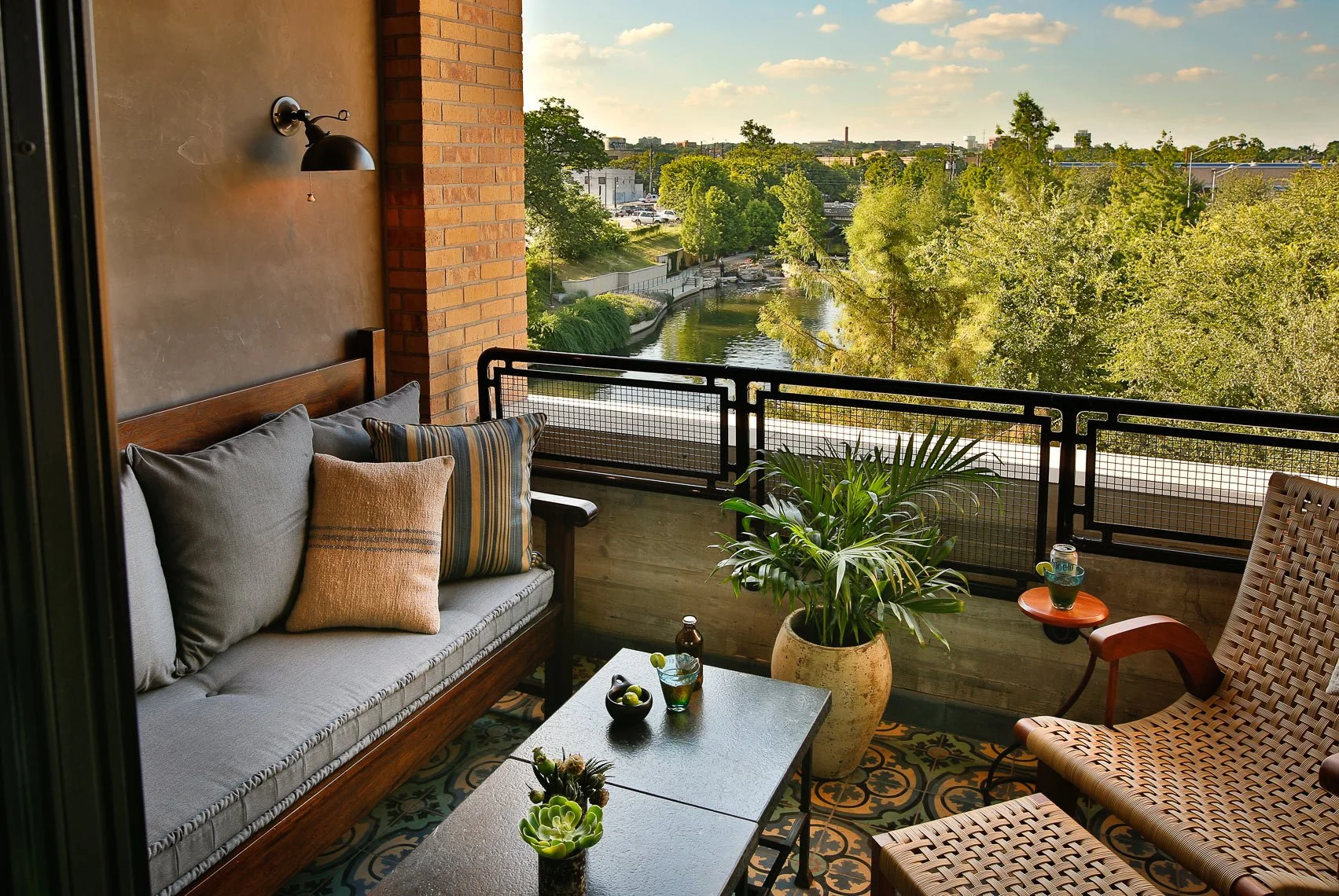 Balcony with outdoor seating, including a sofa with pillows, a woven chair, a potted plant, and a view of a river lined with trees and buildings in the background.