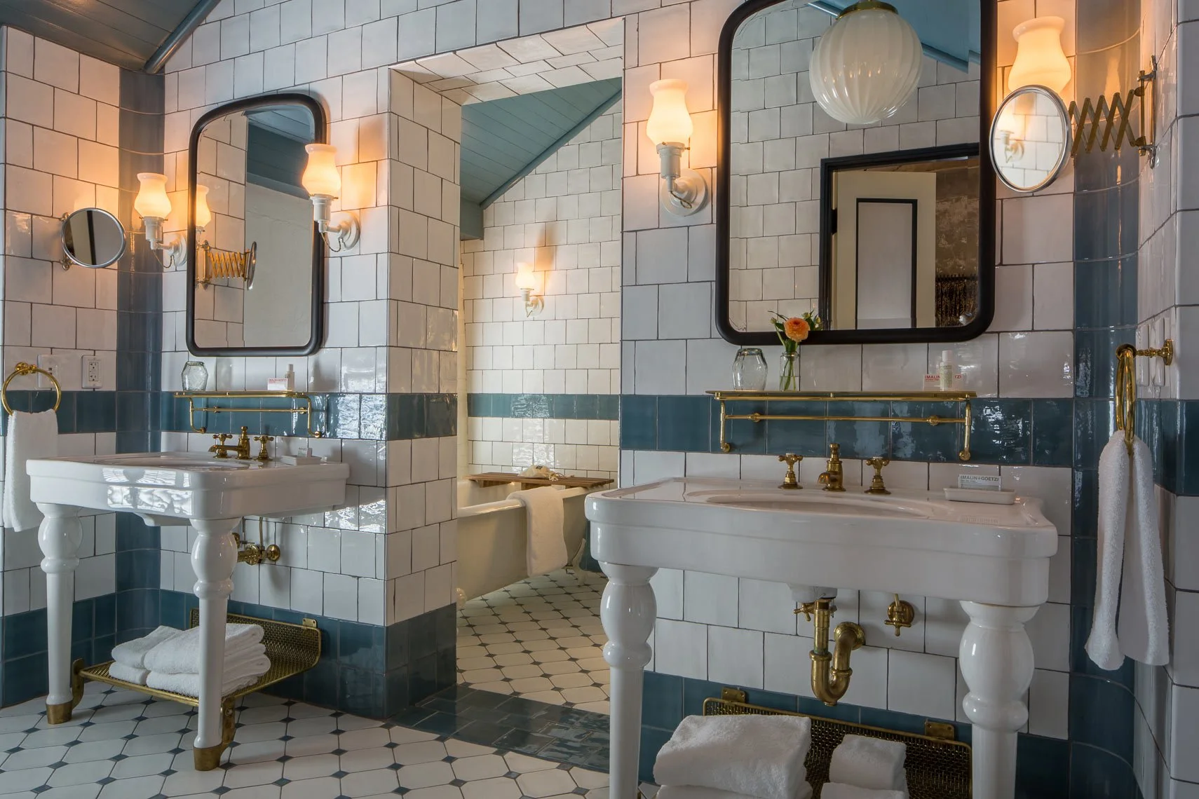 A vintage-style bathroom with two white sinks and large rectangular mirrors, decorated with wall-mounted lights and gold fixtures, featuring a tiled wall with white and blue tiles, and a small bathtub in the background.