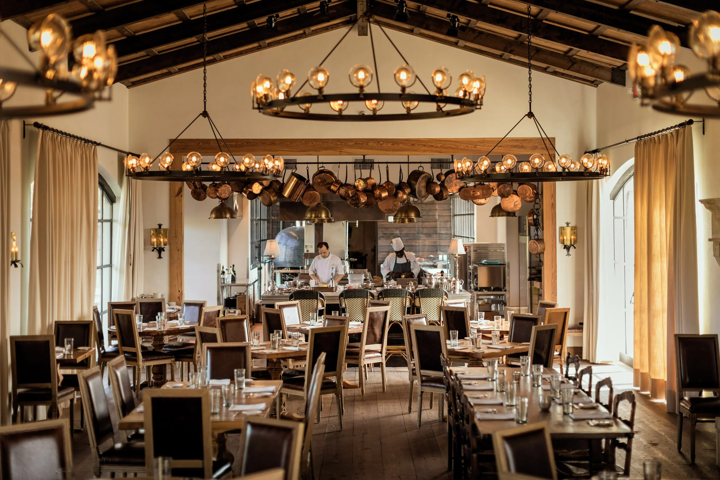 Interior of a restaurant with multiple tables set with glasses, with a high ceiling, hanging pendant lights and chandeliers, beige curtains, and staff preparing food in the background.