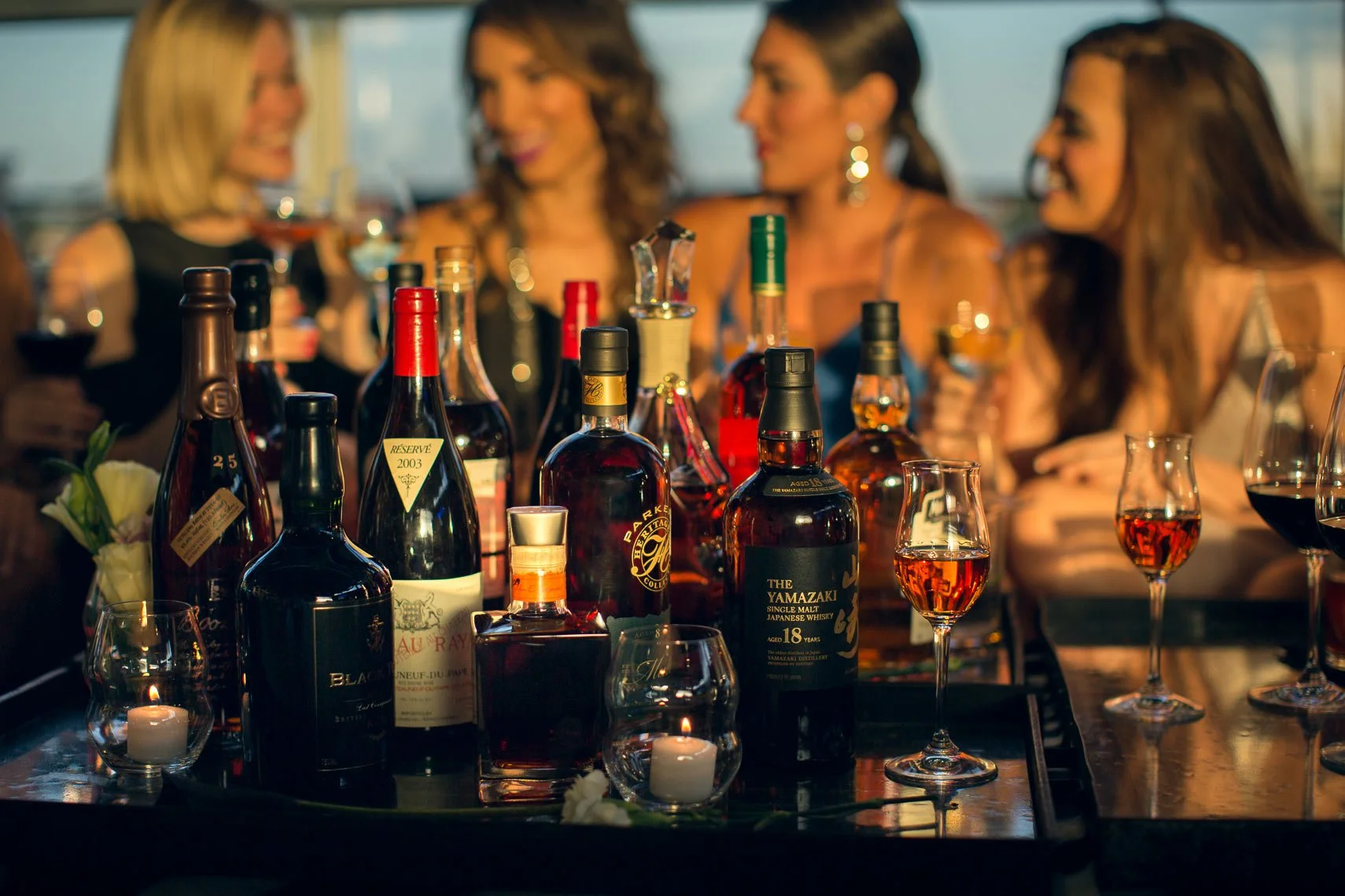Four women socializing at a liquor-filled bar or lounge, with various bottles of whiskey and wine on the table, and candles providing soft lighting.