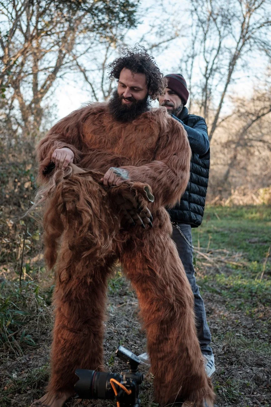 Two men outdoors, one in a large brown furry costume resembling a Bigfoot, the other helping adjust it, with trees in the background.
