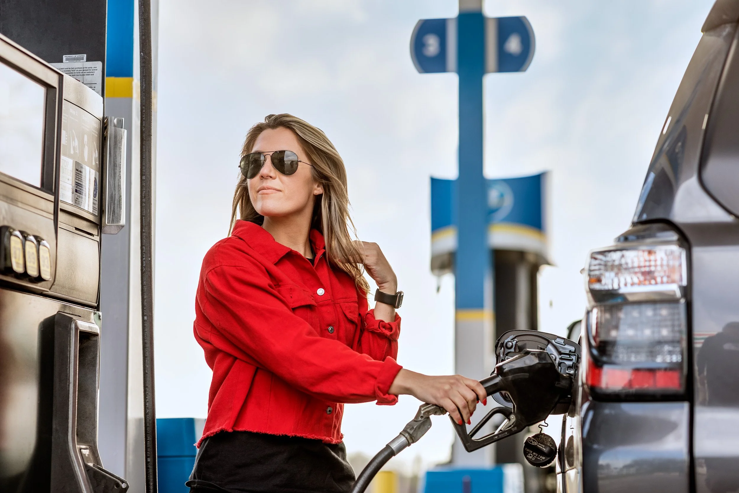 A woman with long hair wearing sunglasses, a red jacket, and black pants, filling up her car with fuel at a gas station.