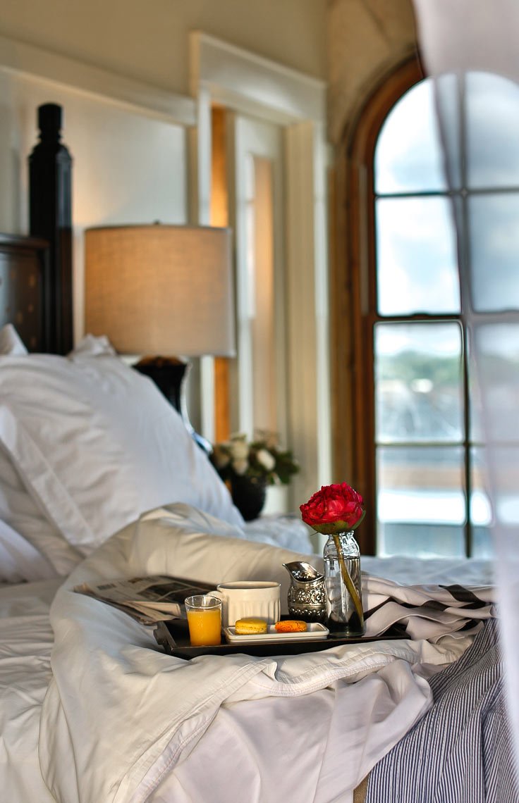 A hotel bed with white sheets, a tray holding a glass of orange juice, a mug, cookies, and a small flower vase with a pink flower, in front of a window with curtains and a lamp.