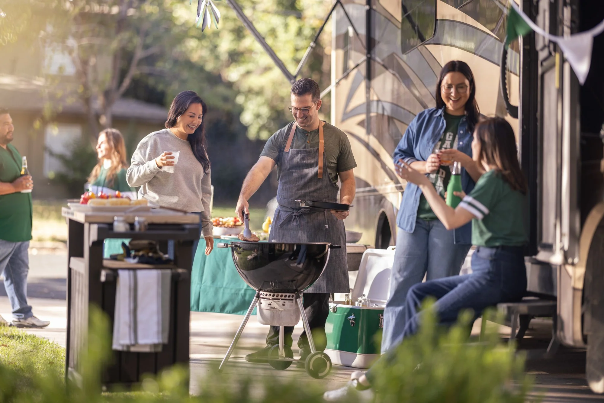 People enjoying an outdoor barbecue party, with a man grilling food and others socializing and drinking drinks.