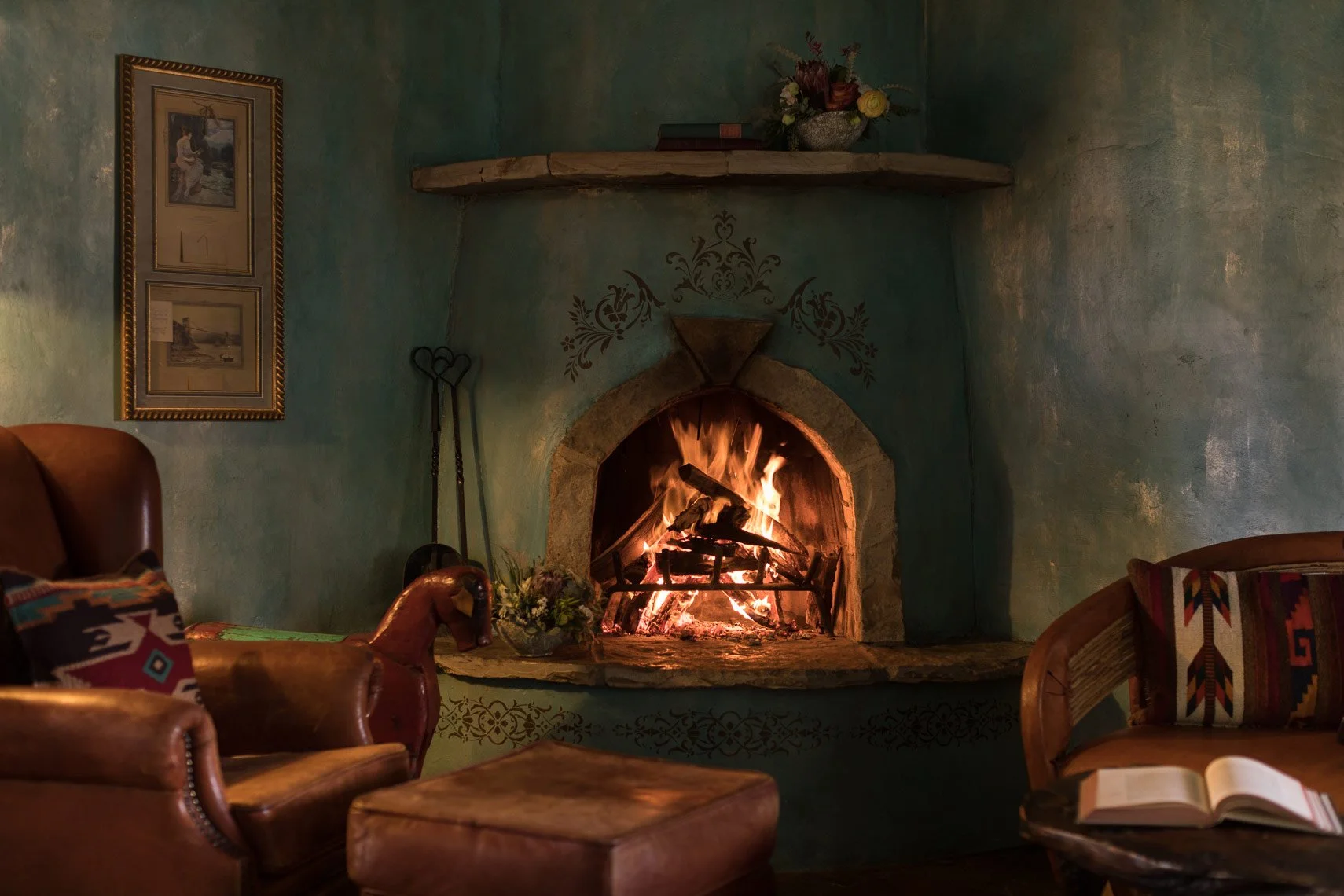 Living room with a lit fireplace, two upholstered armchairs, a wooden coffee table with an open book, framed artwork on teal walls, a floral arrangement on the mantel, and decorative wall art around the fireplace.