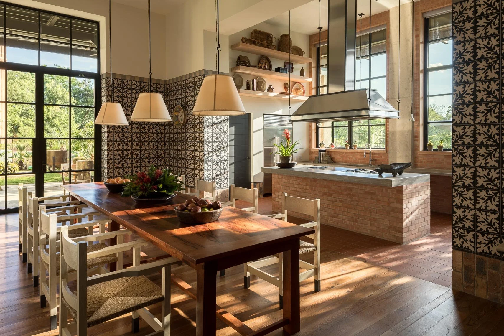 A bright, modern kitchen with large windows, a wooden dining table with chairs, and an island with a brick base and concrete countertop. The room is decorated with hanging pendant lights, shelves with baskets and artifacts, and plants near the window